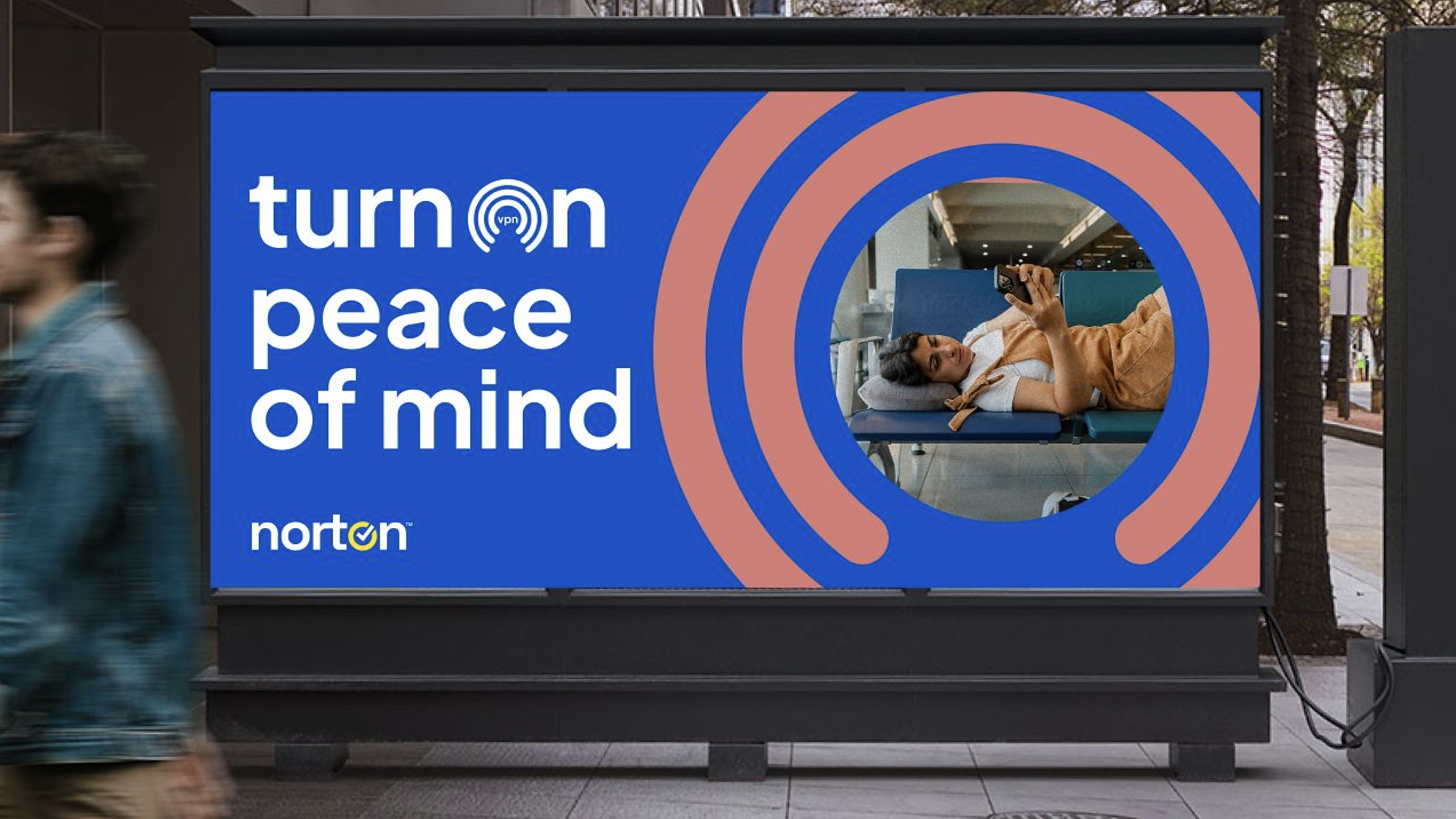 A woman lying on a blue chair in an airport, looking at her phone, behind a large colorful advertisement for Norton VPN, which features the message 'turn on peace of mind' and a circular design.