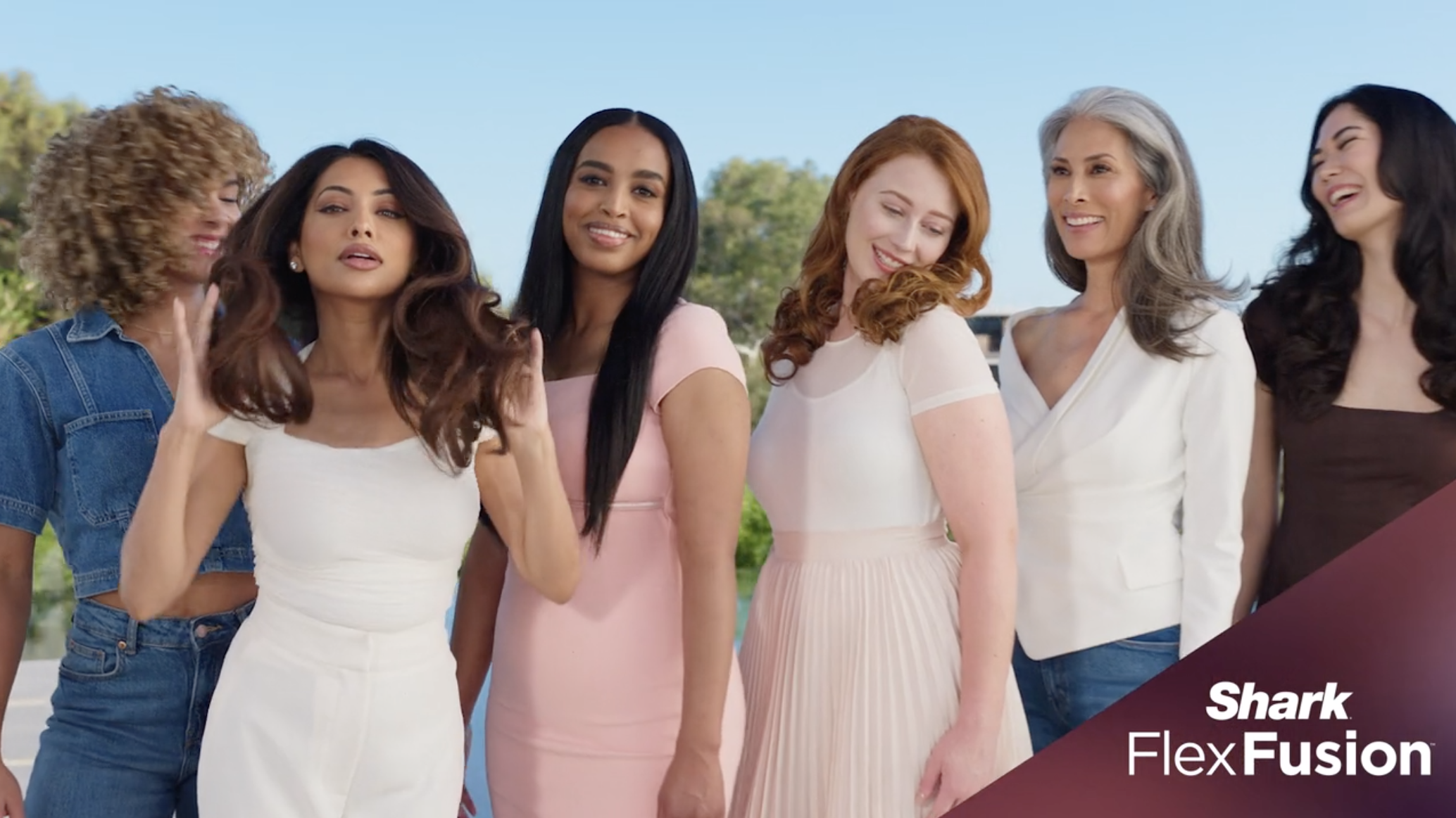 A diverse group of six women standing outdoors, smiling and enjoying each other's company, with a blue sky and greenery in the background.