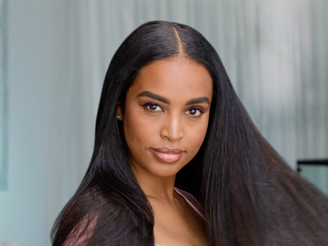 A woman with long, straight black hair, looking at the camera with a slight smile in a light-colored room.