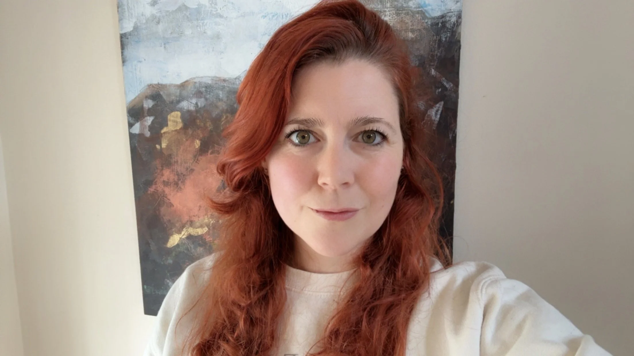 A woman therapist with red hair and green eyes takes a selfie indoors with a wall and artwork behind her.