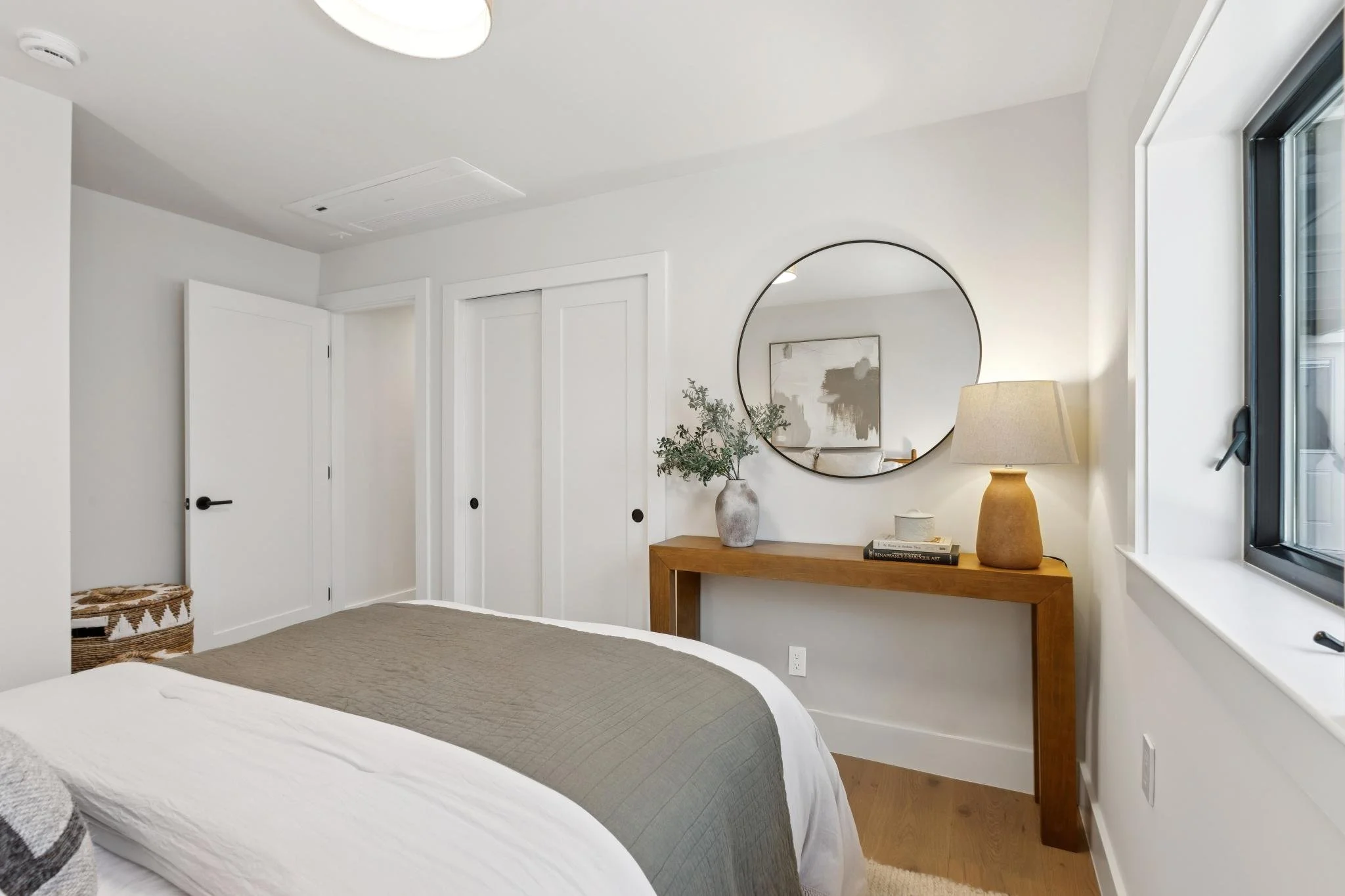 A bedroom with white walls, a window, a wooden dresser with decorative items and a mirror, and part of a bed with gray and white bedding.