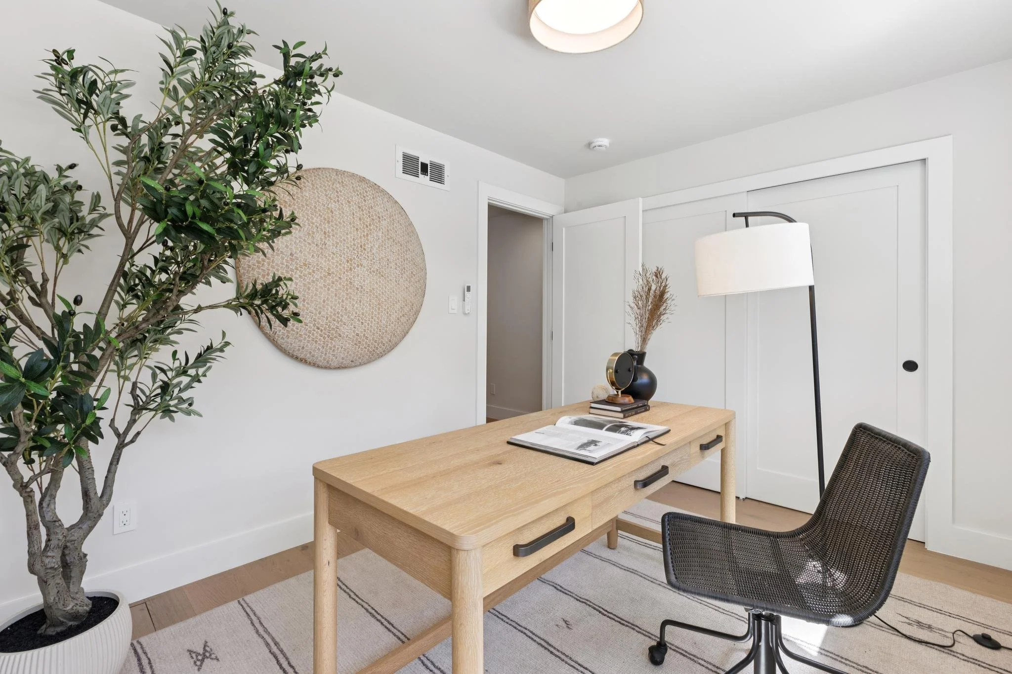 A modern, minimalist home office with a large potted olive tree, a round woven wall decor, a wooden desk with an open magazine, a black chair, a tall floor lamp with a white shade, and a small vase with dried plants. The room has white walls, a door,