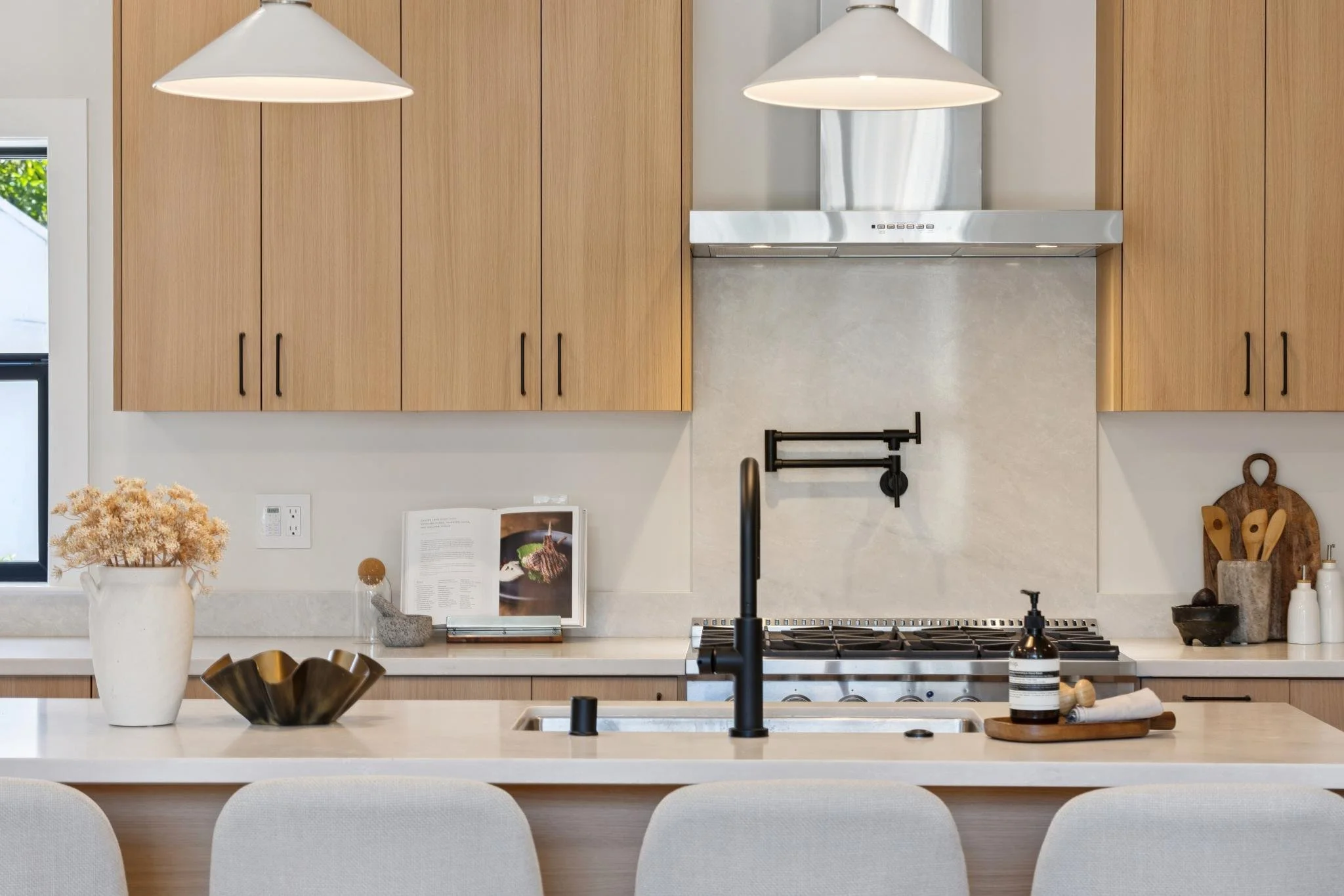 Modern kitchen with light wood cabinets, white countertops, black faucet, black stovetop, and decorative items including a white vase with dried flowers, a black bowl, and a small book.