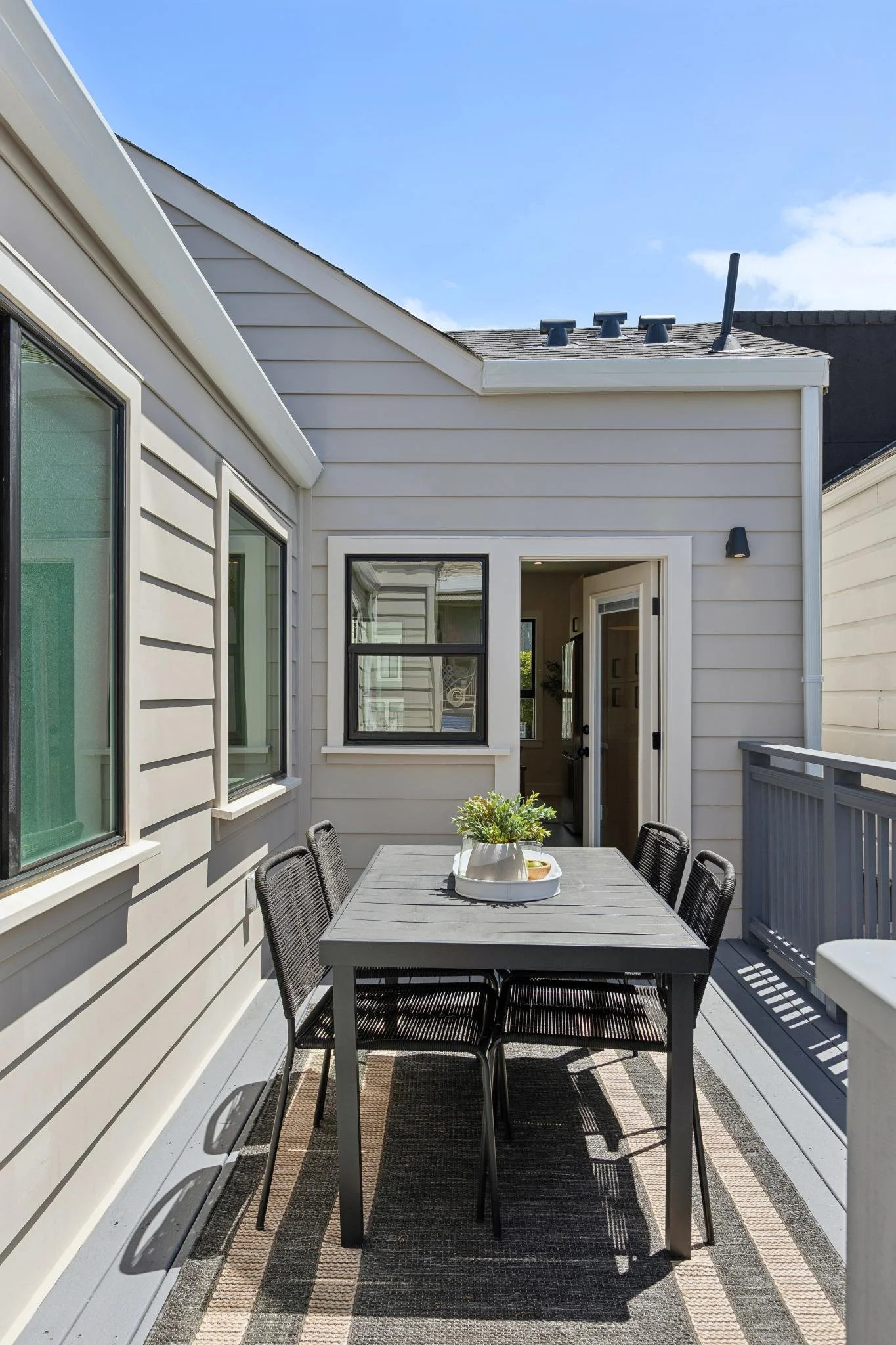 An outdoor balcony with a dark gray dining table, six matching chairs, a plant in a white pot, a gray and beige striped rug, beige siding on the house, and a clear blue sky.