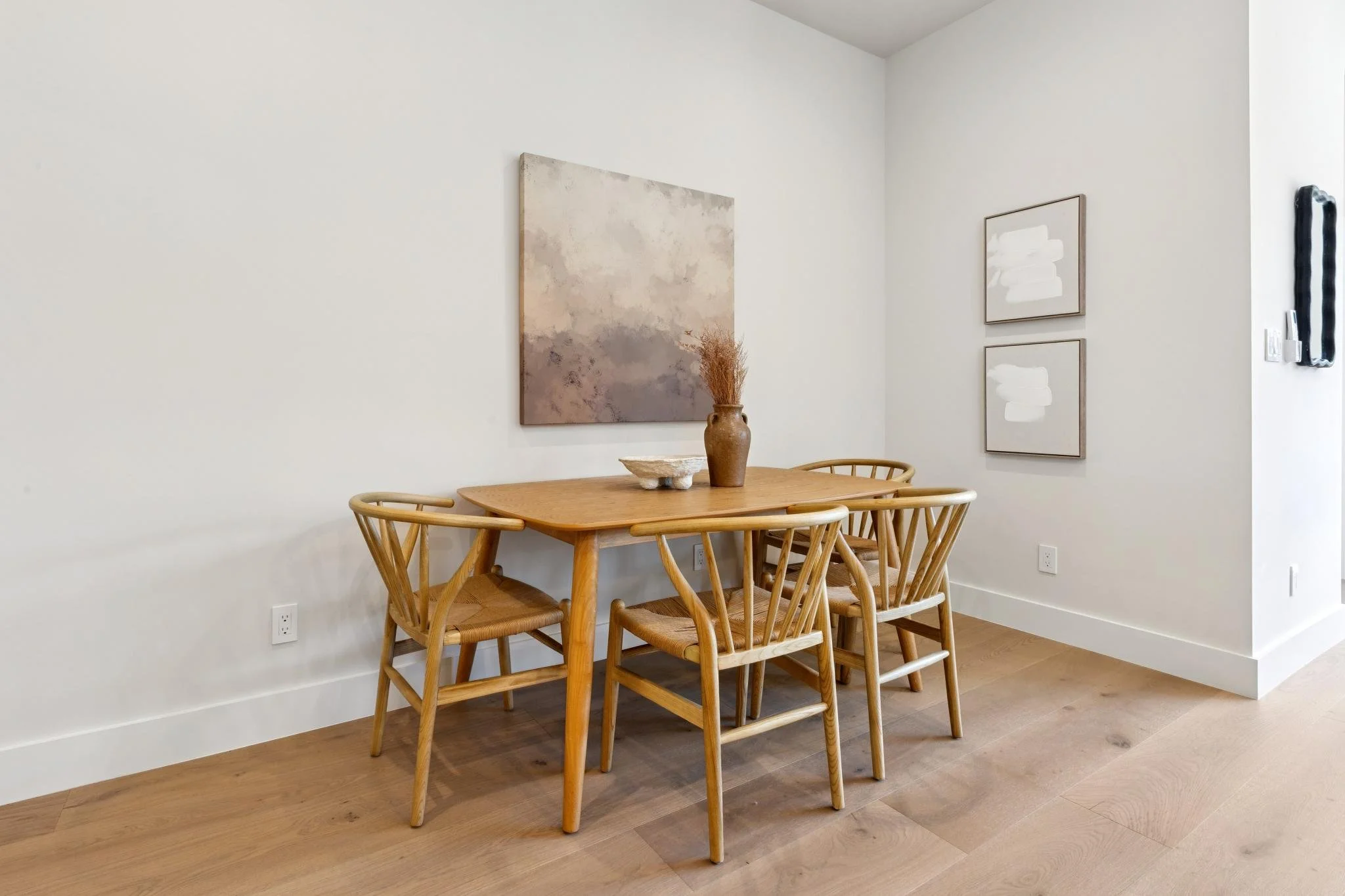 A wooden dining table with six matching chairs in a minimalist room with light-colored walls and art pieces hanging on the walls, including abstract paintings and a decorative mirror.