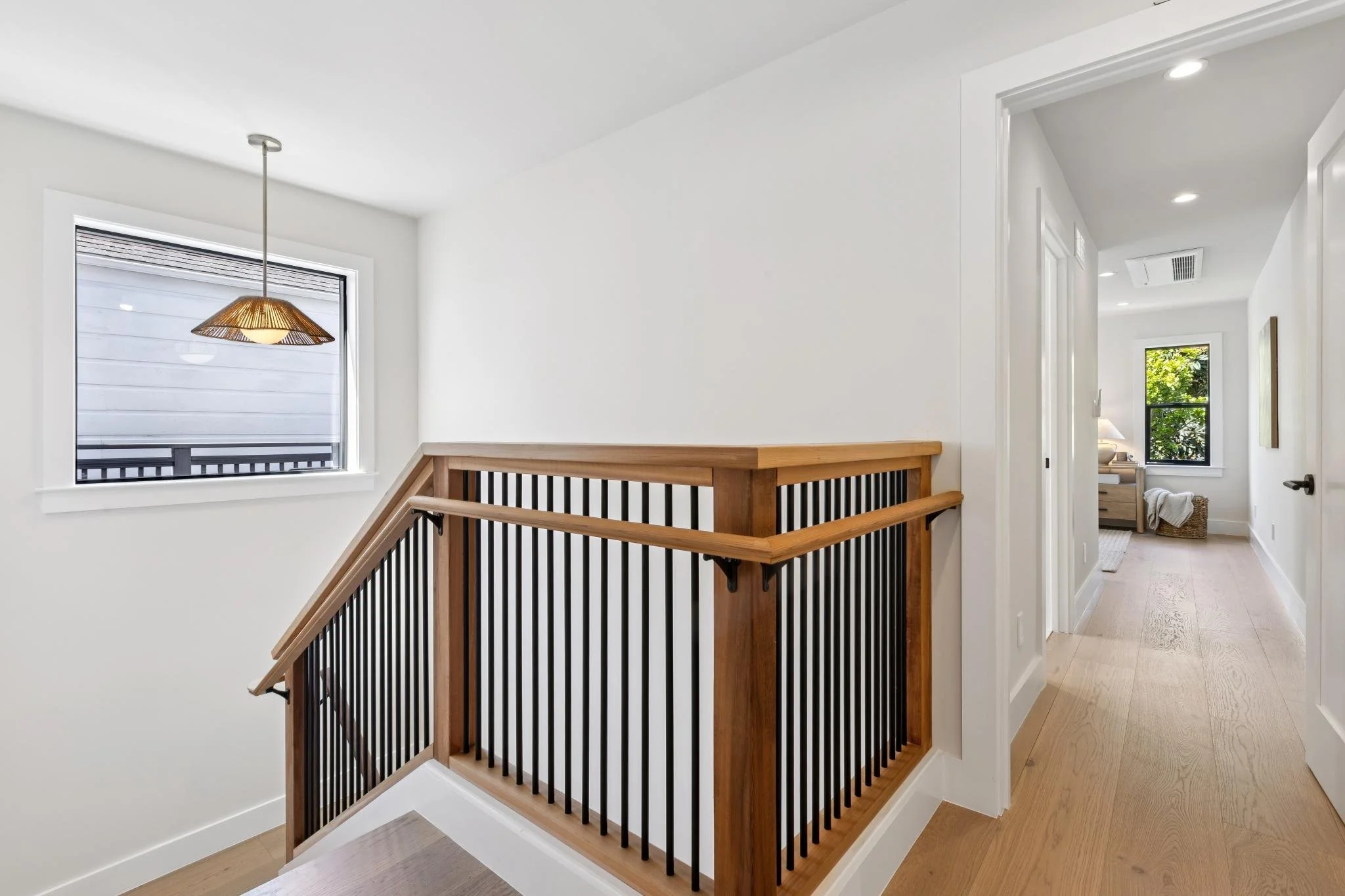 View of a house interior showing a staircase with wooden handrails and black spindles, white walls, multiple windows, and a hallway leading to another room with a window and a light-colored rug.