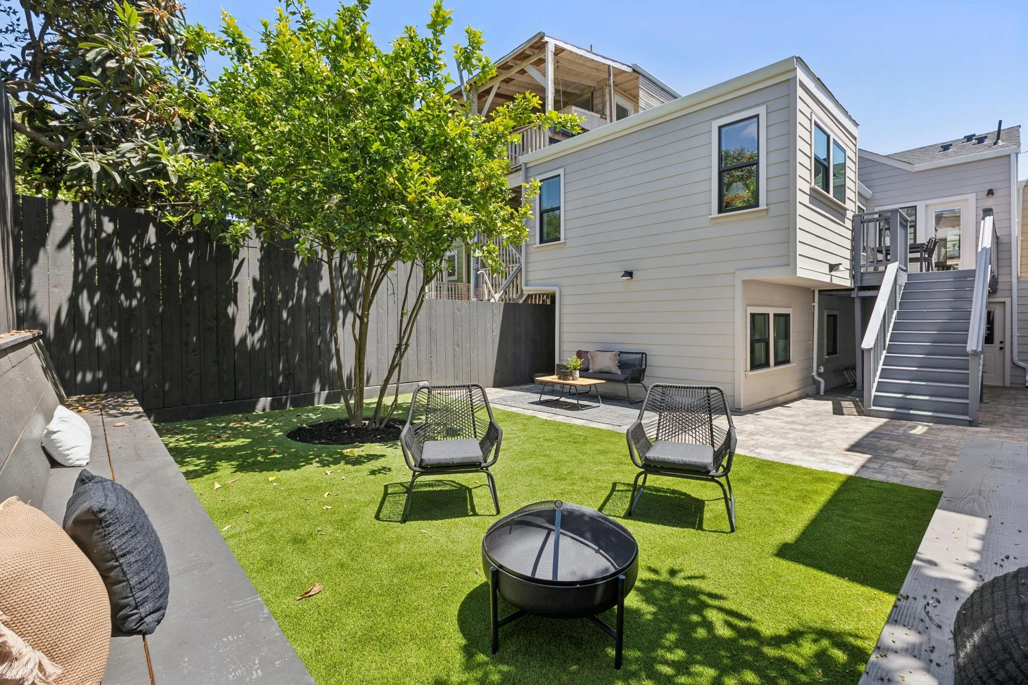 A backyard with green artificial grass, a tree, outdoor furniture including chairs and a bench, a dark fence, and a modern building with multiple levels and stairs, under a clear blue sky.