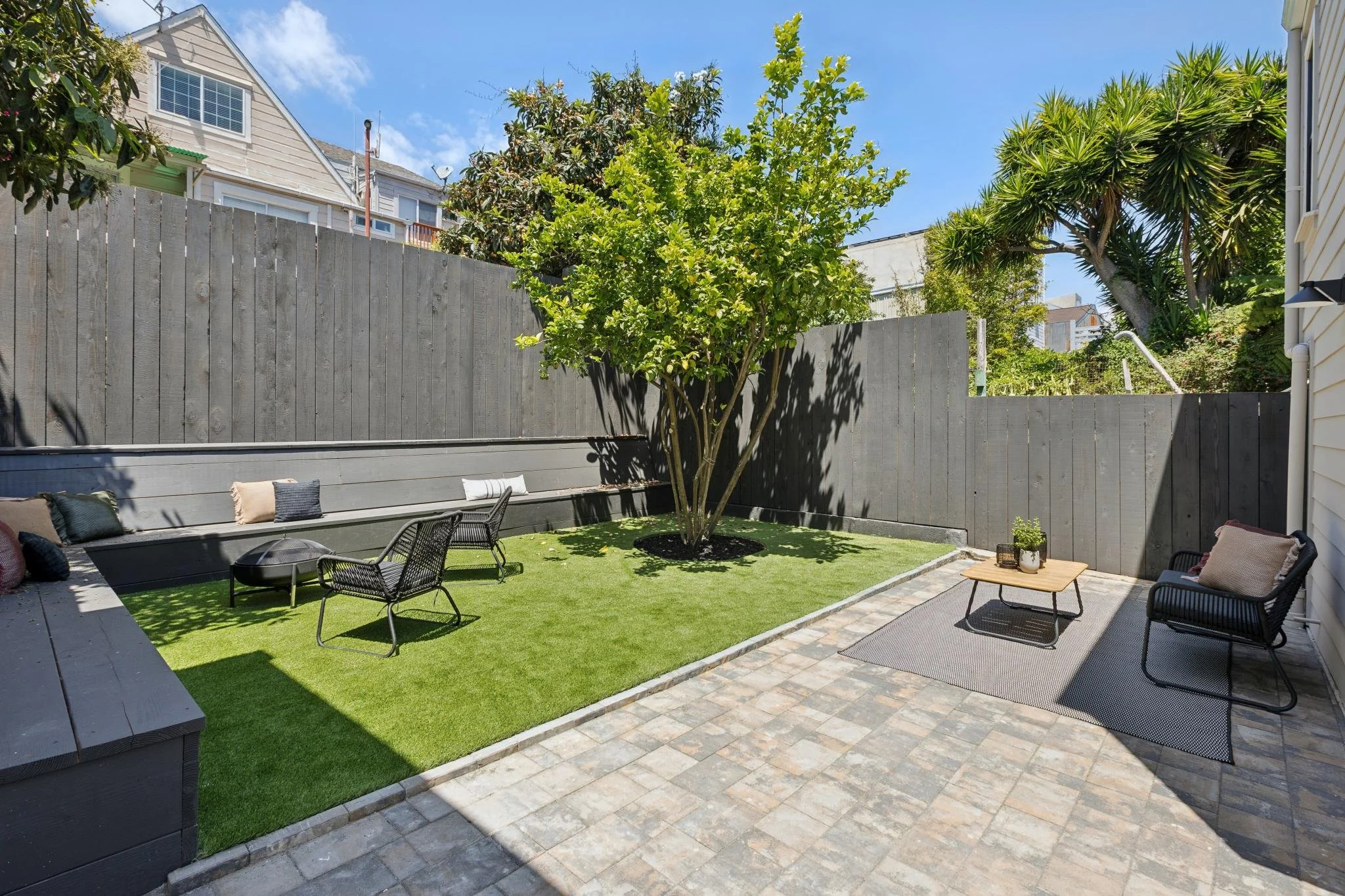 A backyard patio featuring outdoor seating, a tree with lush green foliage, a wooden privacy fence, a small artificial grass area, and a brick walkway with a grey outdoor rug.
