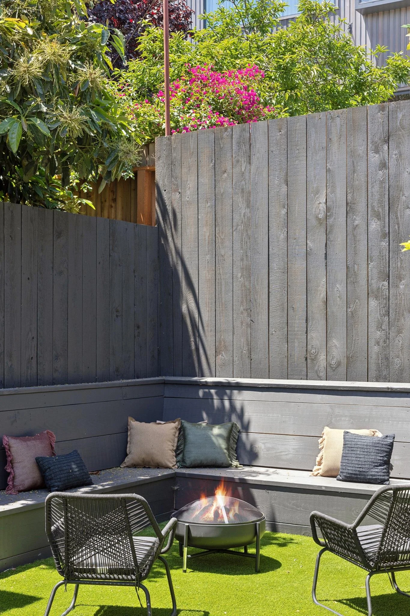 An outdoor patio with a fire pit and black metal chairs on artificial grass, surrounded by a gray wood fence and green plants and flowers.
