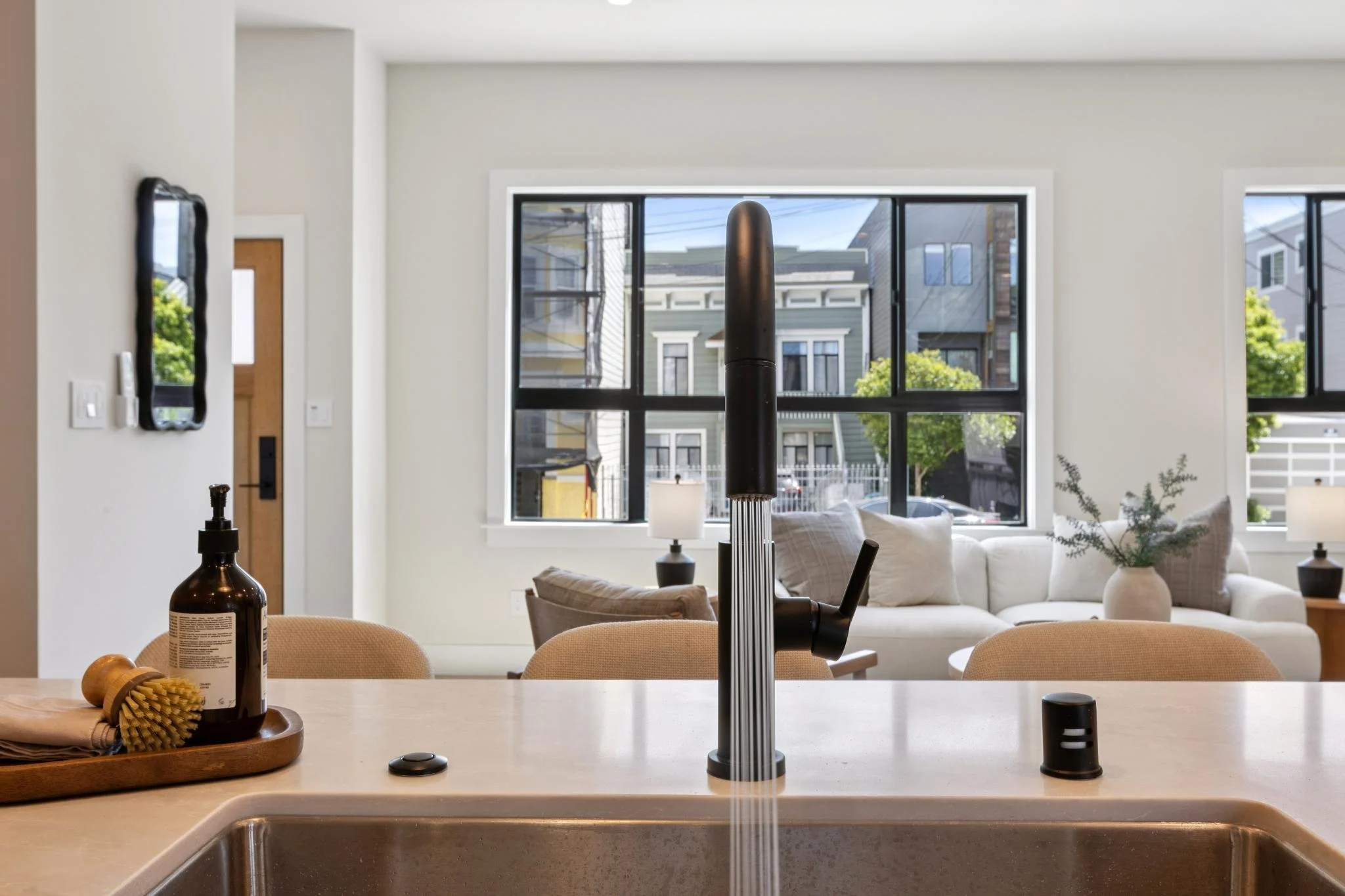 The photo shows a modern kitchen with a white countertop, a sink, and a soap dispenser. In the background, there is a living room with a white sofa, beige chairs, and large windows revealing houses and trees outside.