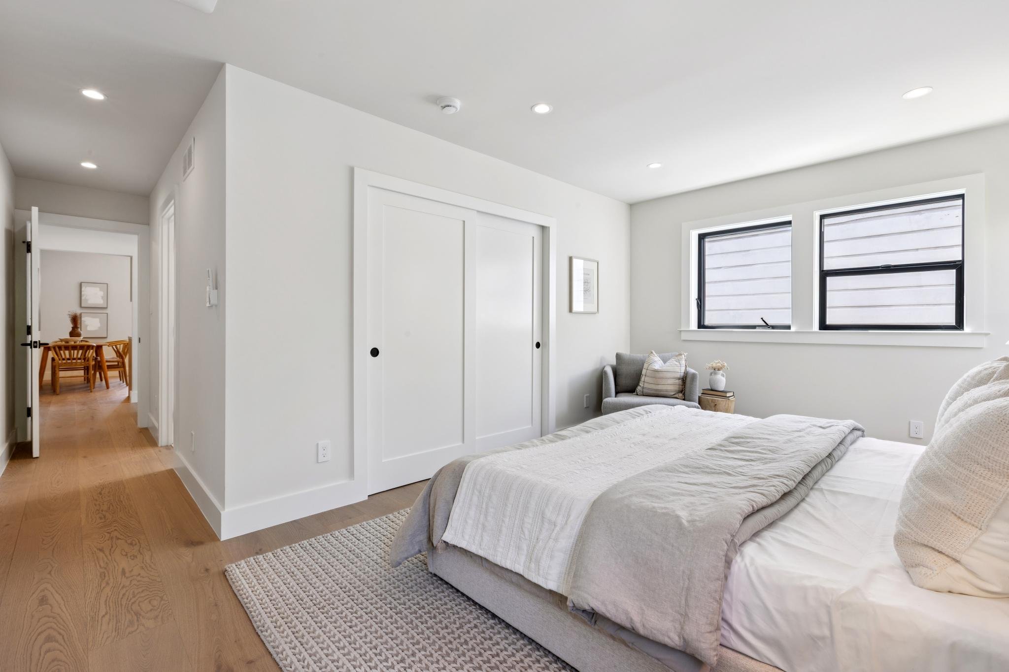 A clean, modern bedroom with white walls, hardwood flooring, and minimalist decor. There is a bed with white bedding, a small gray armchair, and large windows with black frames letting in natural light.