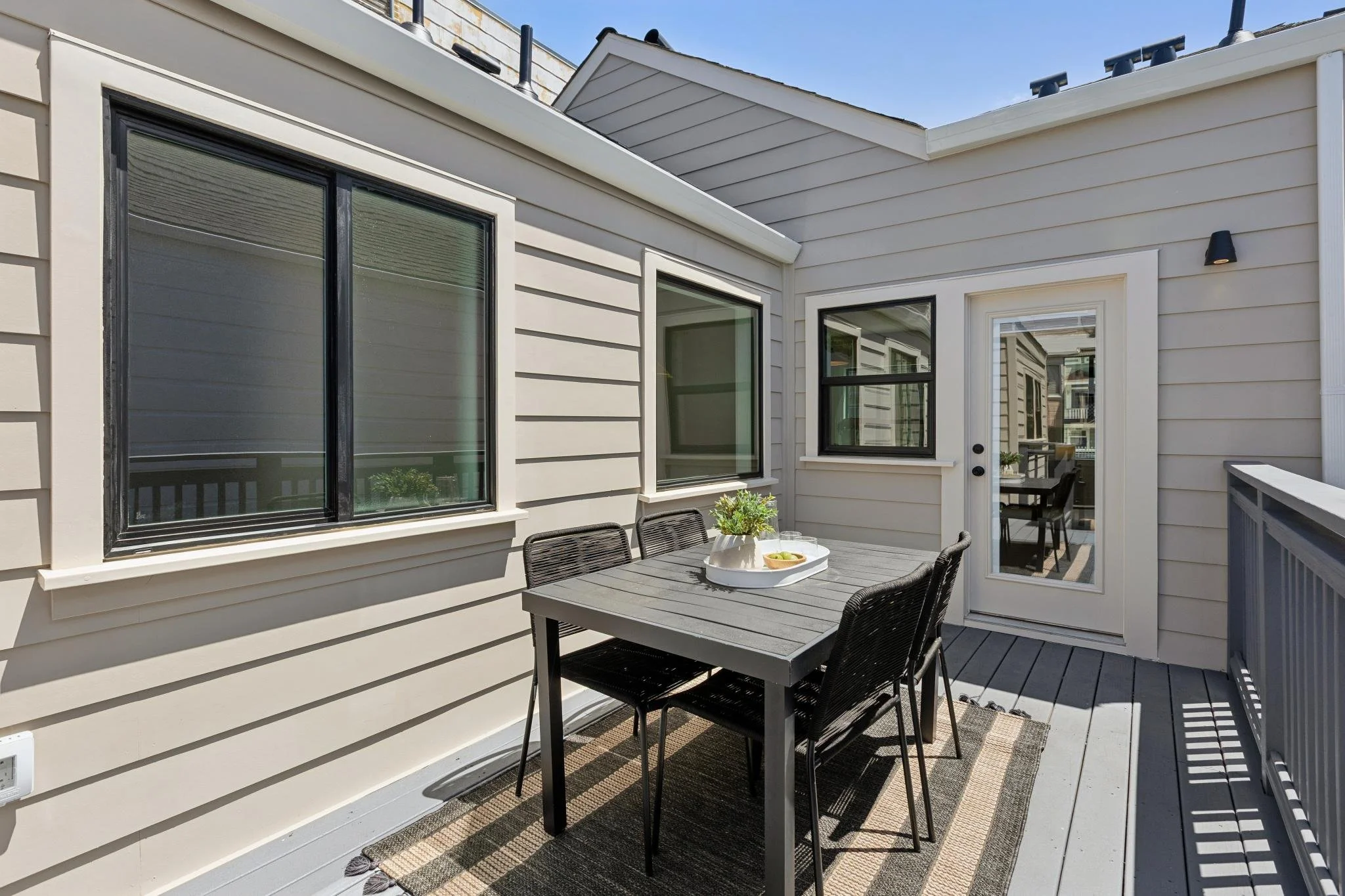 A small outdoor patio area with a gray dining table, four black chairs, a small white tray with a bowl of fruit, and a potted plant, surrounded by beige siding on the house with black-framed windows and a glass door.