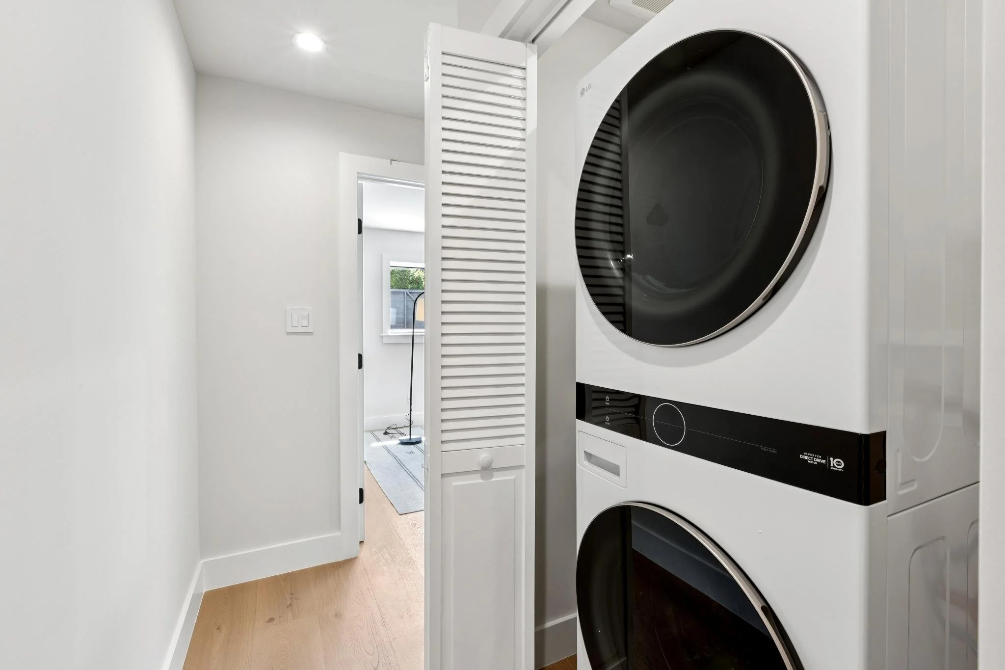 A laundry area with white walls, a stacked LG washer and dryer, a white louvered door, and natural light coming from a window in an adjacent room.