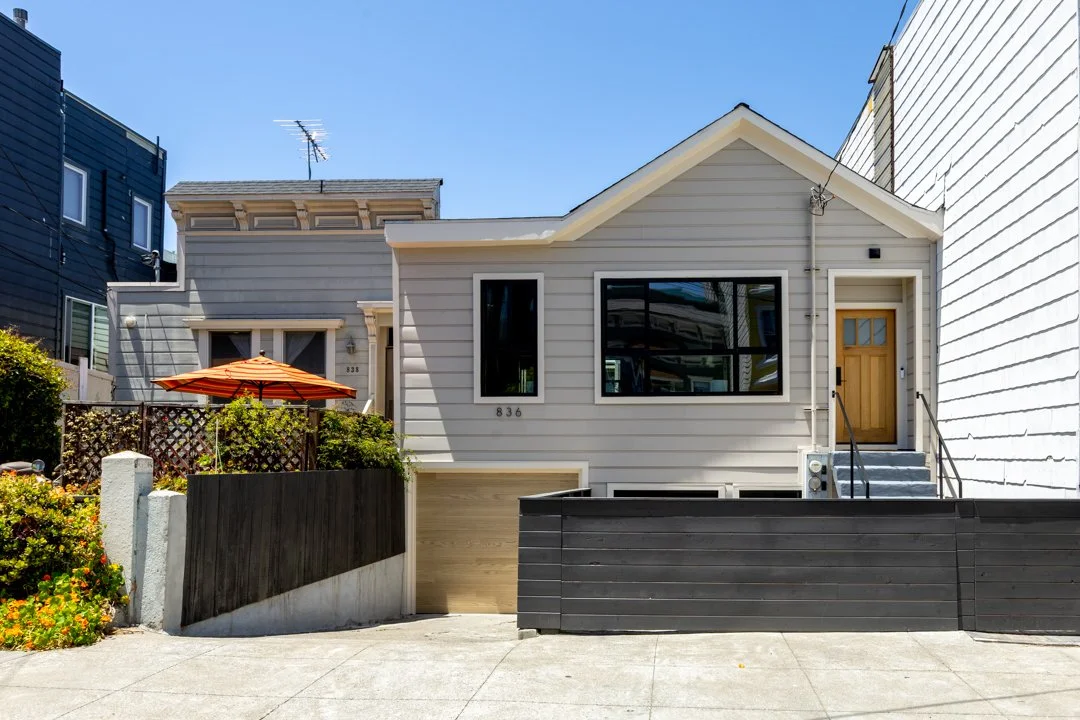 Modern gray house with a wooden front door, black-framed windows, and a small front yard with an orange umbrella and greenery, situated on a sloped street.