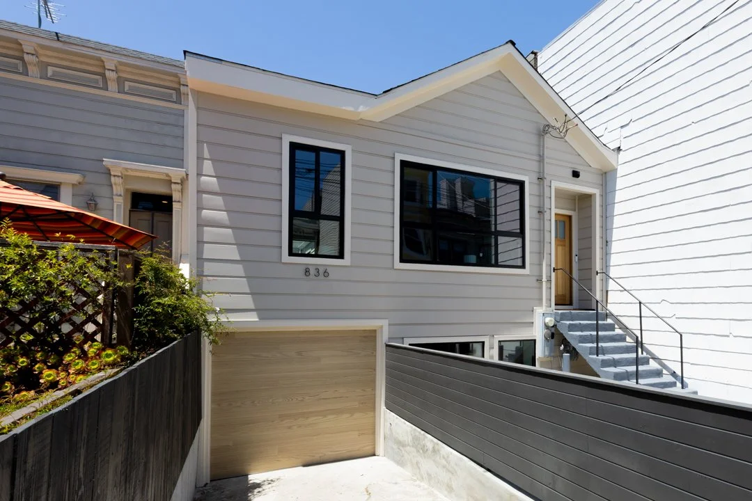 Front view of a modern gray house with black framed windows and a wooden door, accessed by concrete stairs with metal railing, next to a white house with horizontal siding.