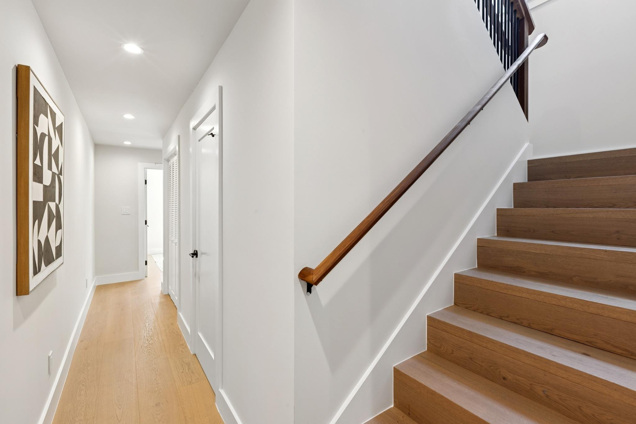 Interior view of a residential hallway featuring wooden stairs with a metal handrail, white walls, and recessed ceiling lights.