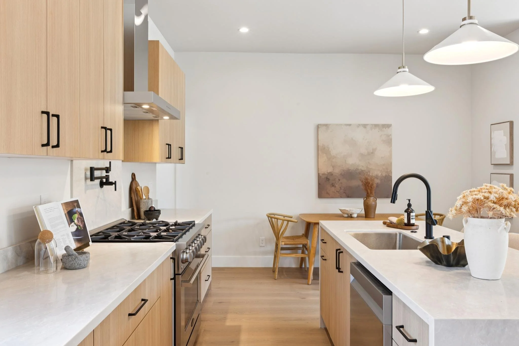 Modern kitchen with light wood cabinets, white countertops, black hardware, gas stove, and kitchen island. Decor includes a framed cookbook, stone mortar, wooden cutting board, vase with dried flowers, and black soap dispenser. There are two wooden c