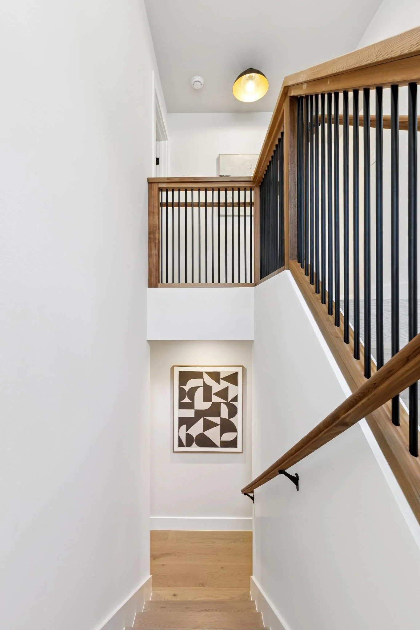 Interior view of a staircase with wooden handrail and black metal balusters leading to the upper floor, white walls, and a framed black and white geometric art piece on the wall at the bottom of the staircase. The ceiling has a black and yellow light