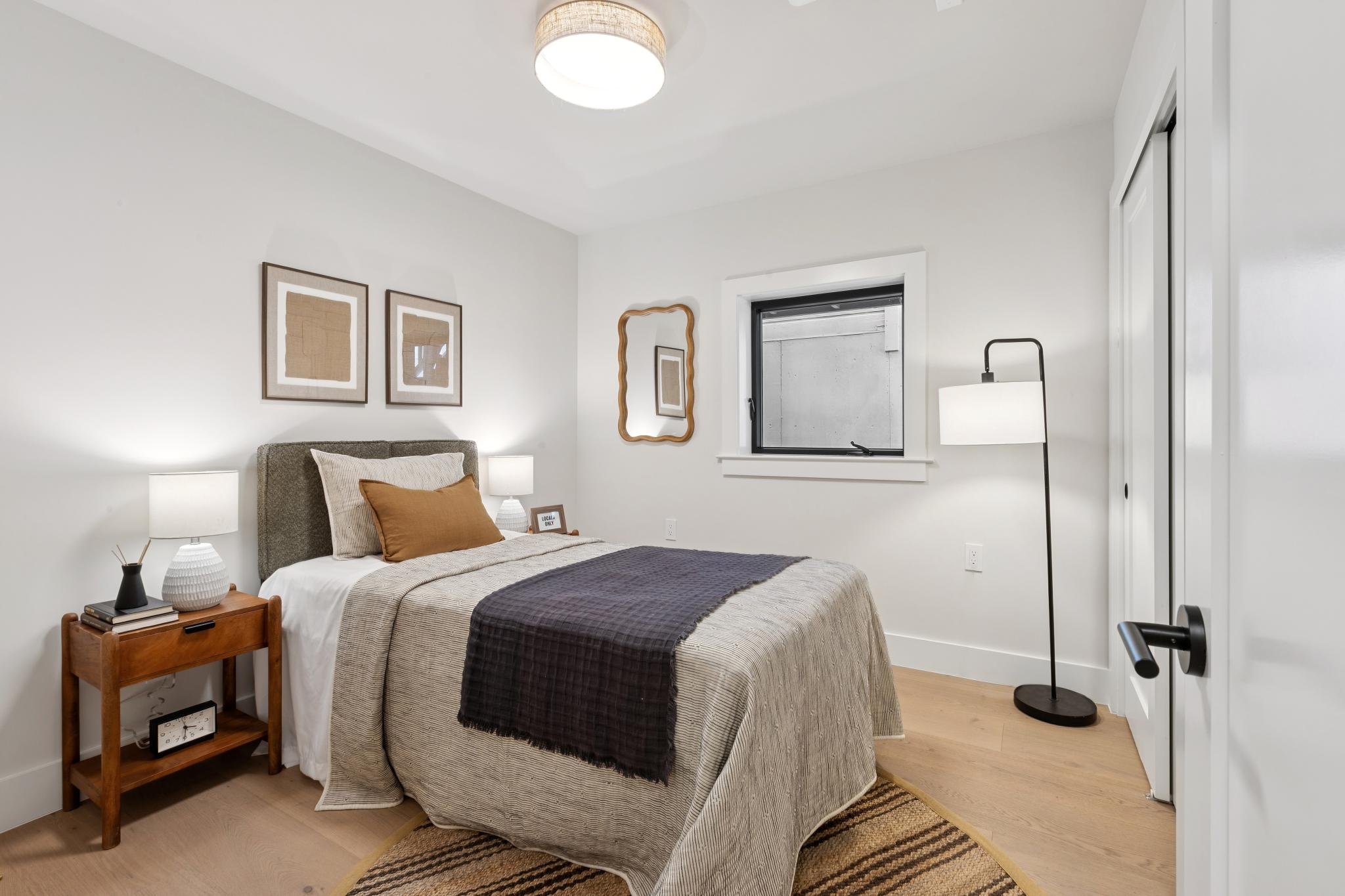 A cozy bedroom with a bed that has a beige and white striped bedspread, brown and beige pillows, and a dark gray blanket. The nightstand beside the bed holds a lamp, books, and a clock. There are two framed artworks above the bed, a mirror on the wal