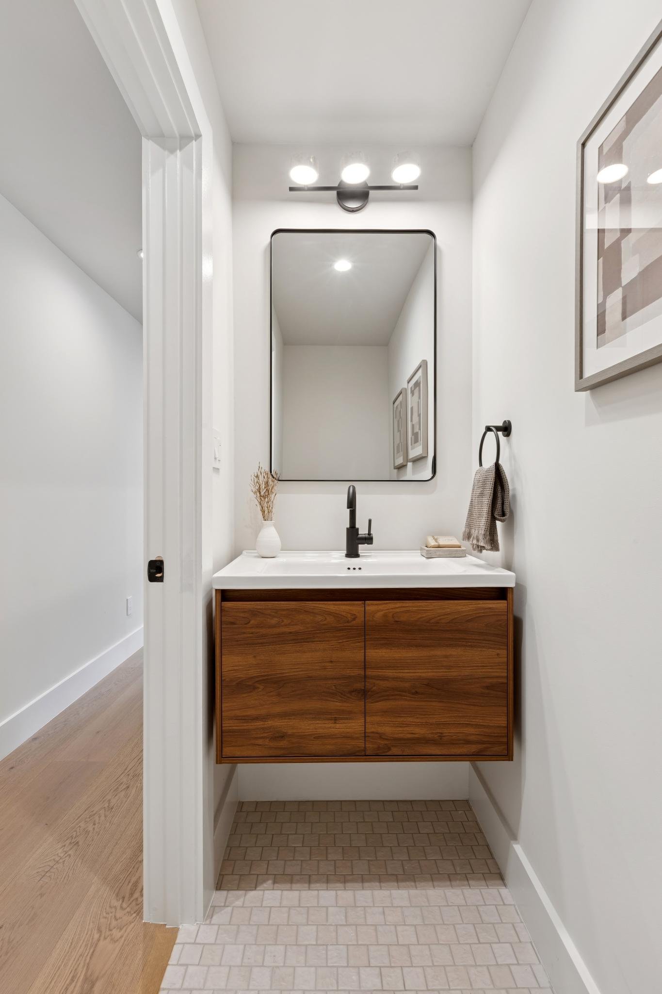 Modern bathroom with a white vanity, a rectangular mirror, and abstract artwork on the white walls. Towel hanging on a black ring holder and a small bar of soap on the counter. Recessed lighting and beige floor tiles.