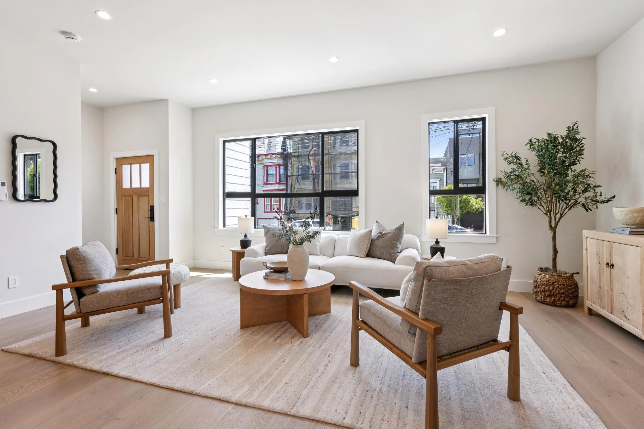 A bright, modern living room with white walls, large windows, and light wooden flooring. There is a white sofa with throw pillows, two wooden armchairs with beige cushions, two small tables with lamps, a potted tree, and a mirror on the wall.