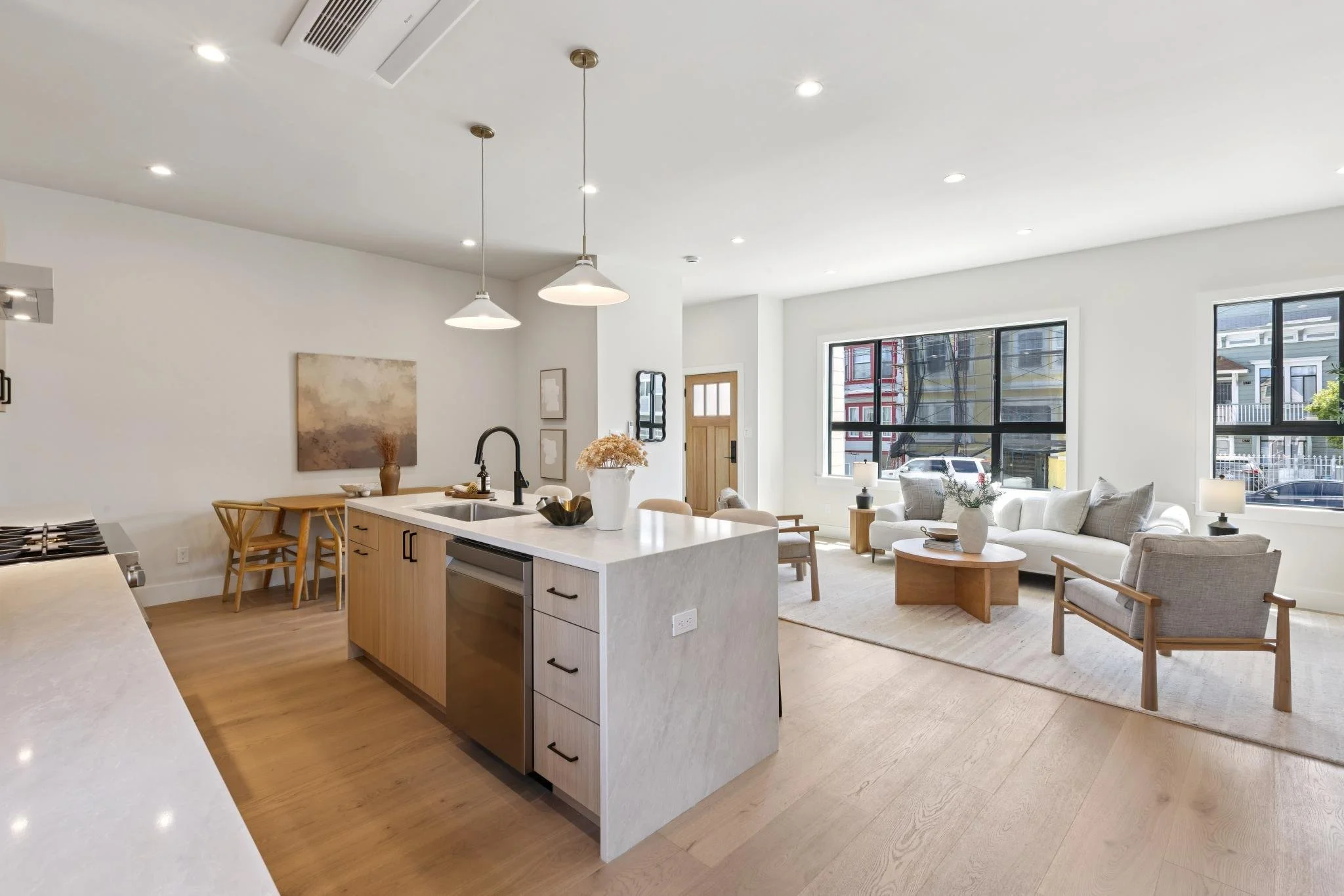 Open-concept living room and kitchen with white walls and large windows showing neighborhood view. The kitchen has a white island with a black faucet and wooden cabinets. The living area has a beige sofa, armchair, round wooden coffee table, and neut
