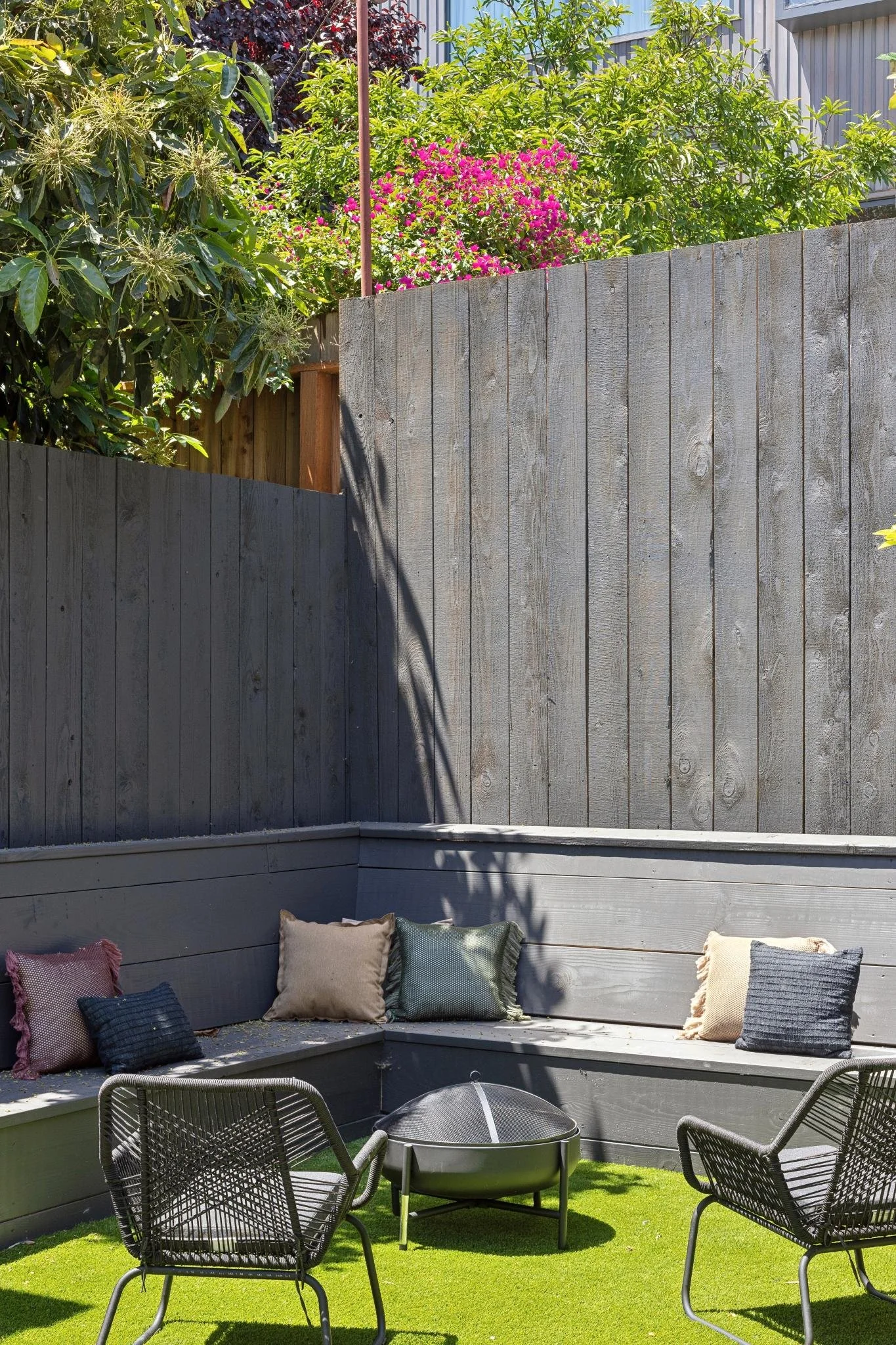 An outdoor patio area with a gray wooden fence, pillows on a built-in bench, and black metal chairs surrounding a fire pit, with green artificial grass ground.