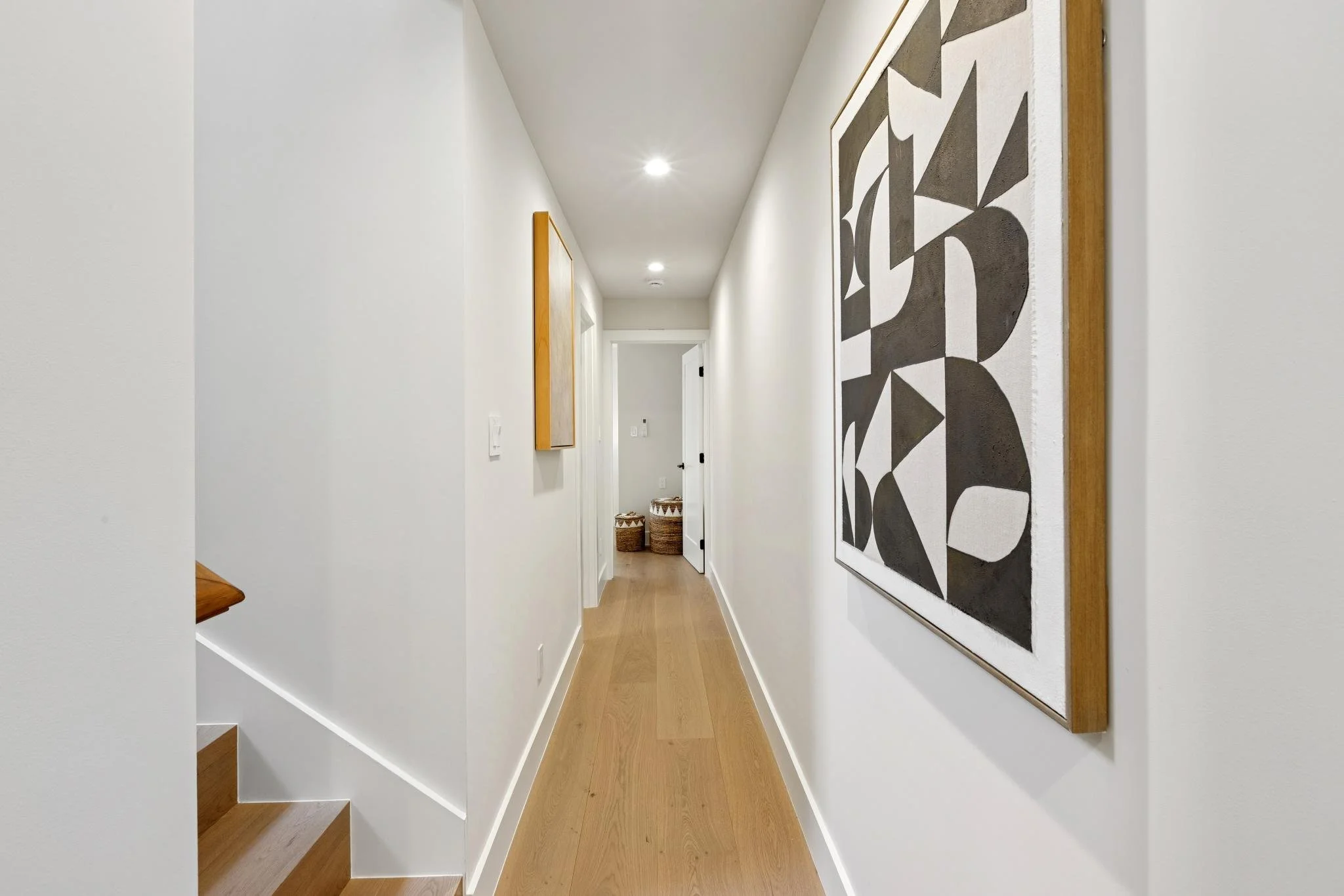 A hallway in a modern home with white walls, wooden flooring, and abstract black and white artwork on the wall. There are baskets at the end of the hallway.