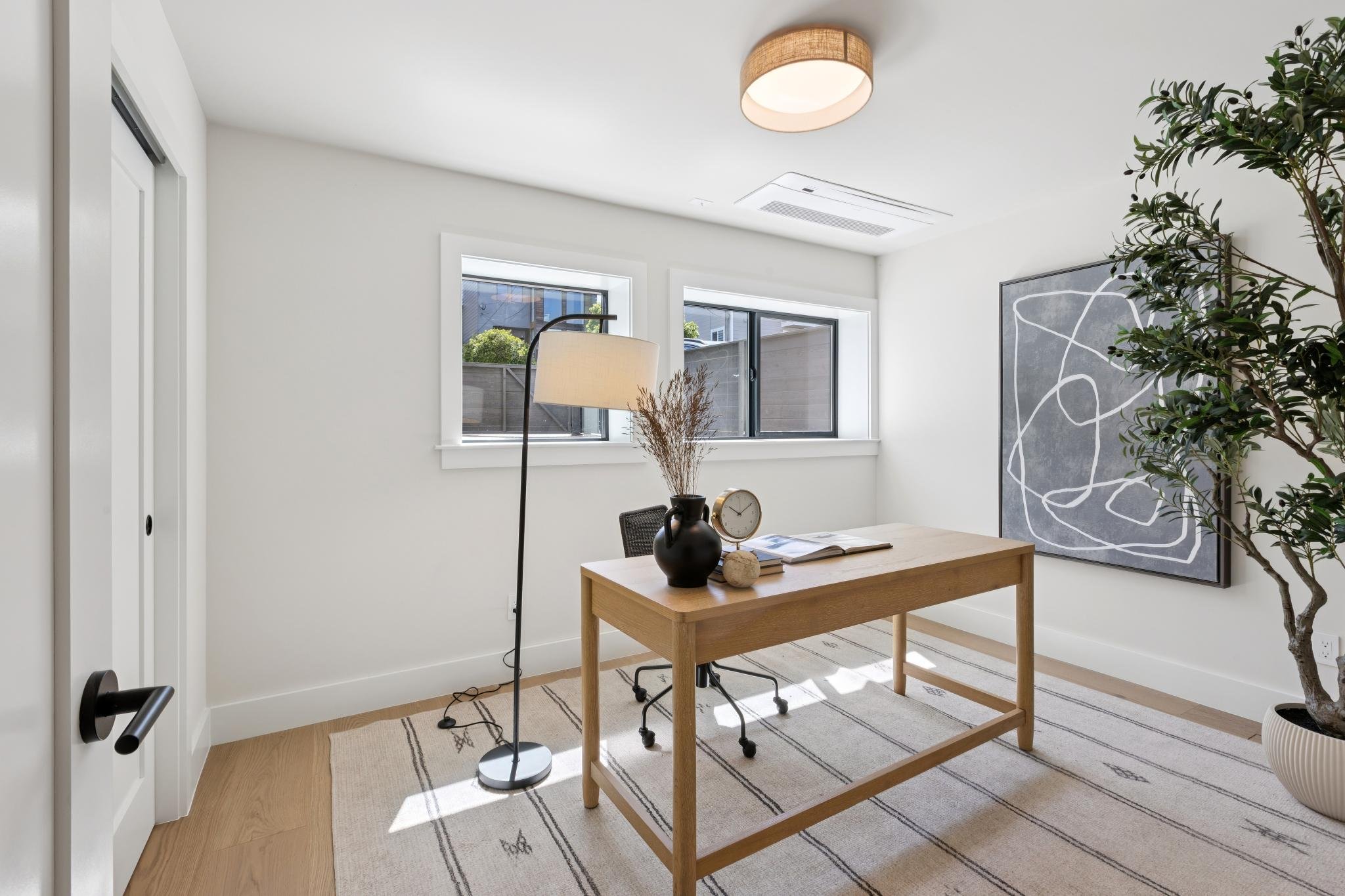 A bright, minimalist home office with a wooden table, black chair, potted plant, abstract artwork, and a tall floor lamp near two windows.