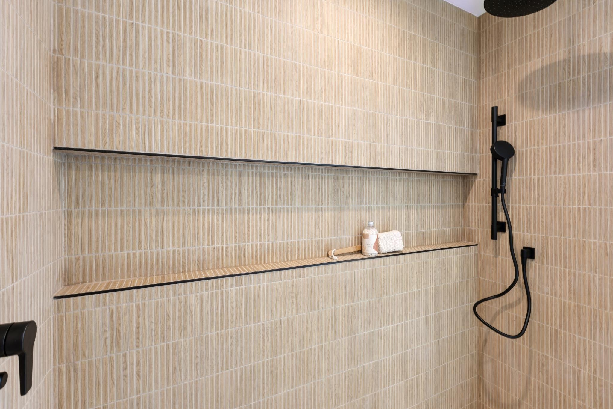 Modern shower with beige rectangular tiles, black fixtures, a handheld showerhead, a shelf with a soap bottle, a towel, and a black wall hook.