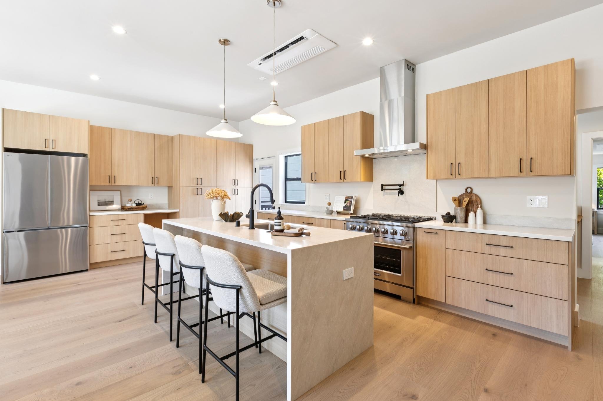 A modern kitchen with wooden cabinets, stainless steel appliances, a kitchen island with a sink, white chairs, and pendant lighting.