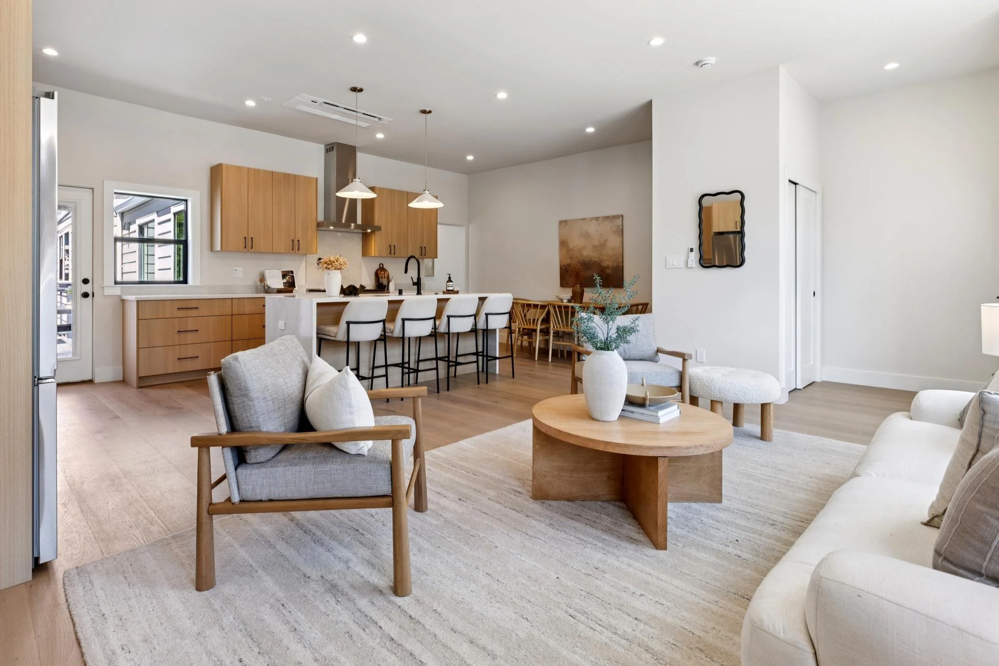 Open concept living room with beige sofa, armchair, and wooden coffee table, adjacent to a kitchen with wooden cabinets, white countertops, island with barstools, and modern lighting, decorated with neutral tones and minimalistic decor.