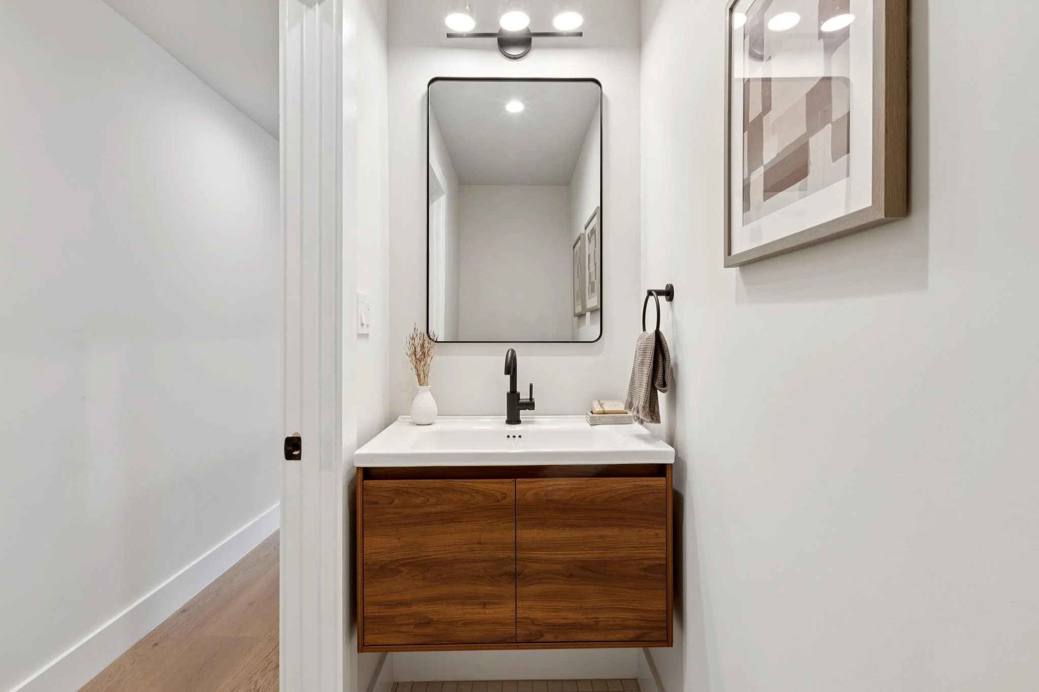 Modern bathroom with white walls, a wooden vanity with a white sink, black faucet, large wall mirror, and minimalist decor including a vase with dried grass and a framed abstract art piece.