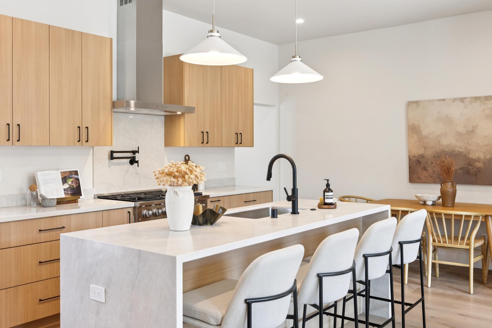 Modern kitchen with light wood cabinets, white marble island, black fixtures, and hanging white pendant lights.