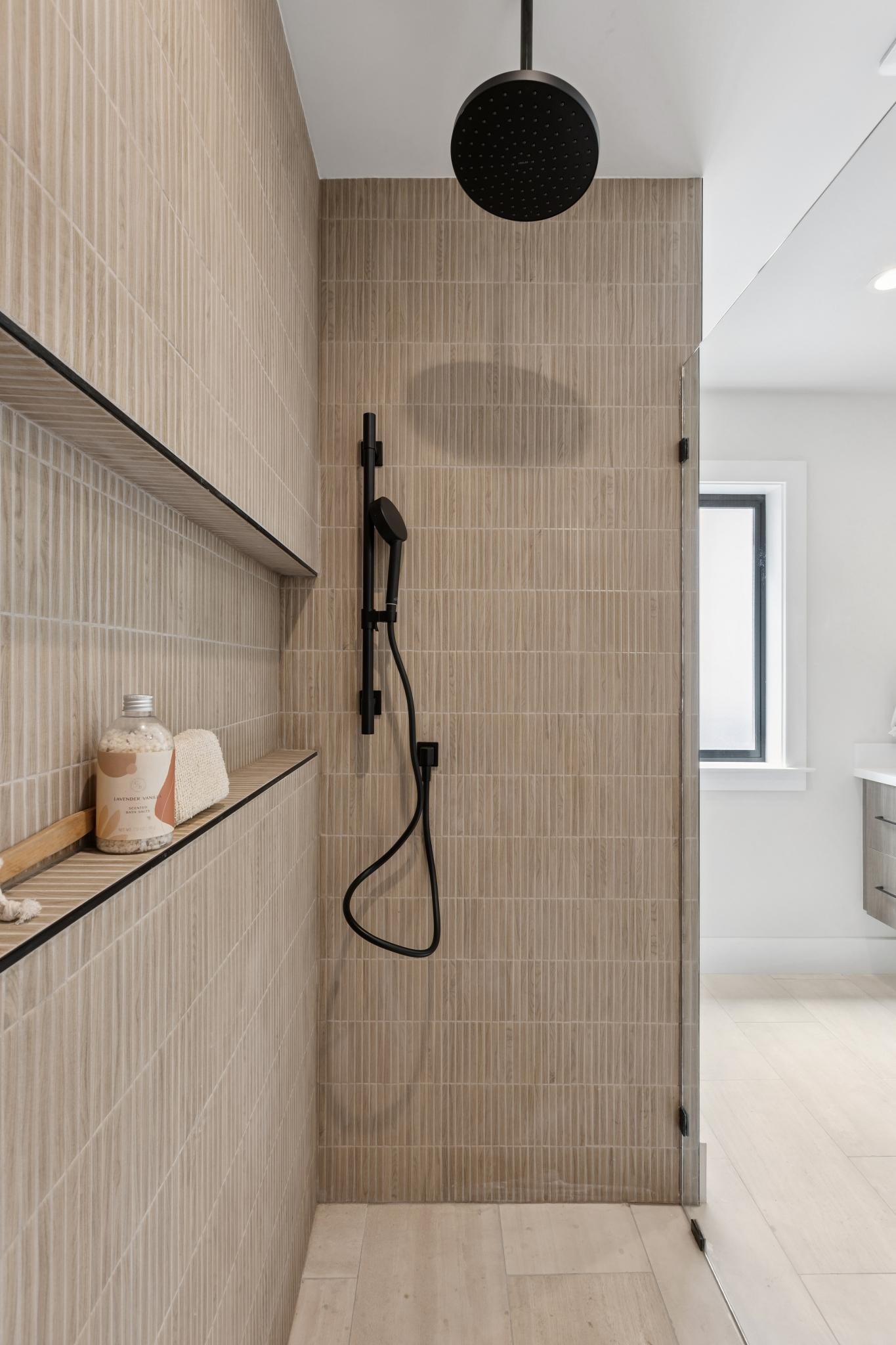 Modern bathroom with a shower area featuring a round black showerhead, a black handheld shower, beige textured wall tiles, a small shelf with a bottle and towel, a large window, and a glass shower door.