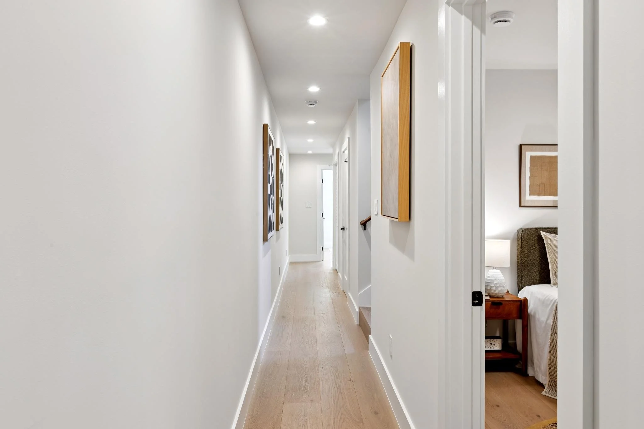 A bright, modern hallway with white walls, recessed lighting, light wood flooring, and framed artwork. Partial view of a bedroom with a bed, nightstand, and a table lamp.