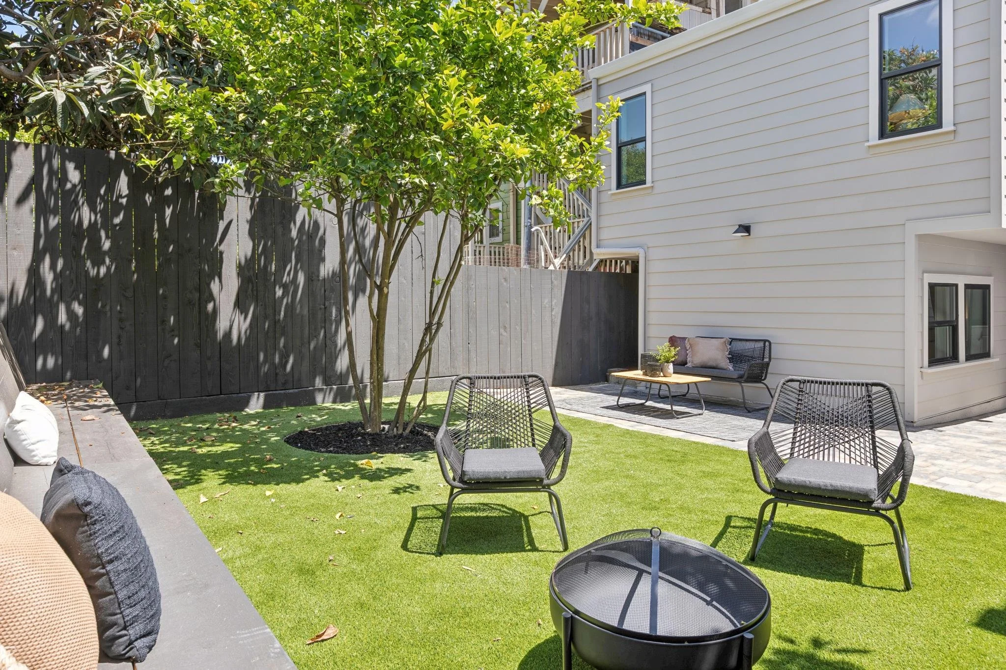 A backyard patio with outdoor seating, a tree, a black fence, and a white house with multiple windows.