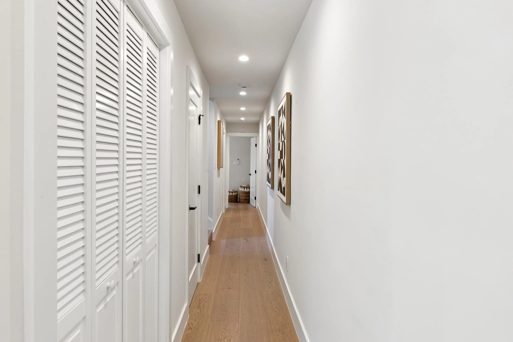 Bright hallway with white walls and ceiling, hardwood flooring, and a series of closed white louvered closet doors on the left. Decor includes framed artwork on the right wall and woven baskets at the end of the hall.