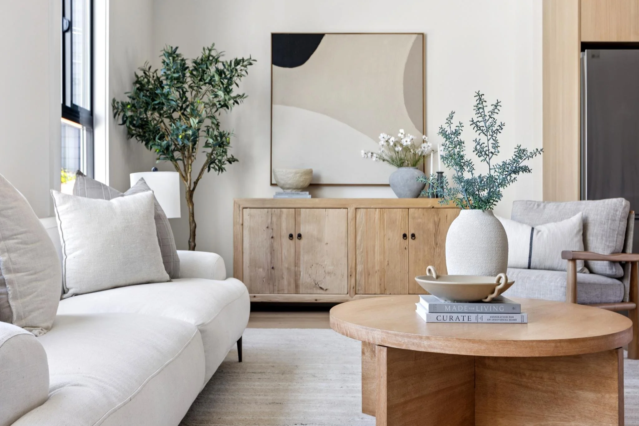 A living room corner decorated with a large green indoor plant, a framed abstract artwork, ceramic vases with white flowers, and neutral-colored sofas and pillows on a light-colored rug.