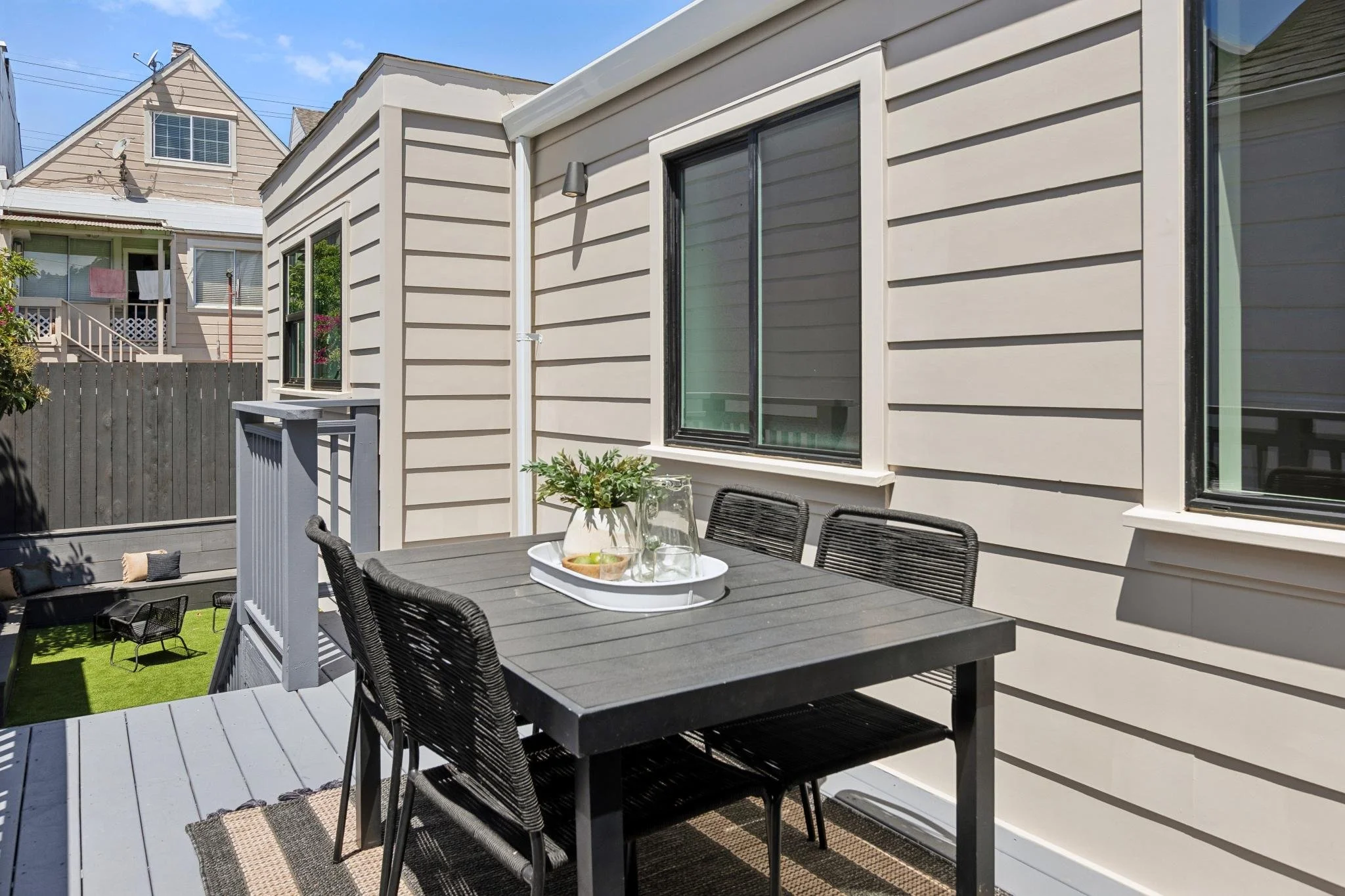 An outdoor patio with a black dining table and chairs, a gray railing, and a house with beige siding and large windows. There are plants and a small grassy area in the background, and neighboring houses are visible. The sky is blue with some clouds.
