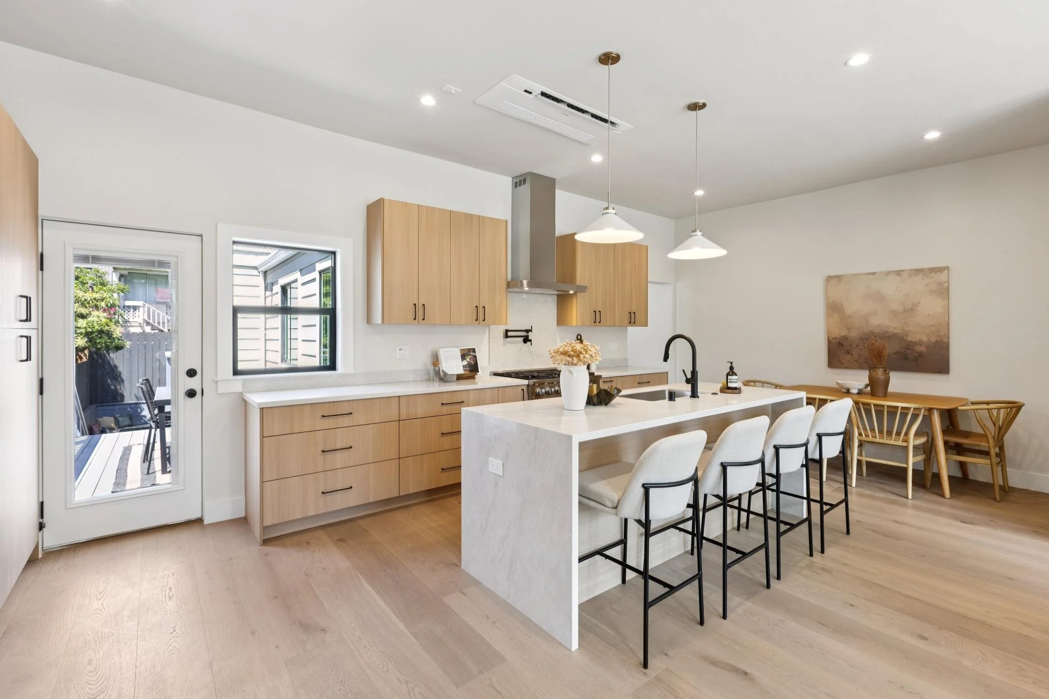 Modern kitchen and dining area with light wood cabinets, white island with seating, black faucet, and pendant lighting, with a sliding glass door leading outside.