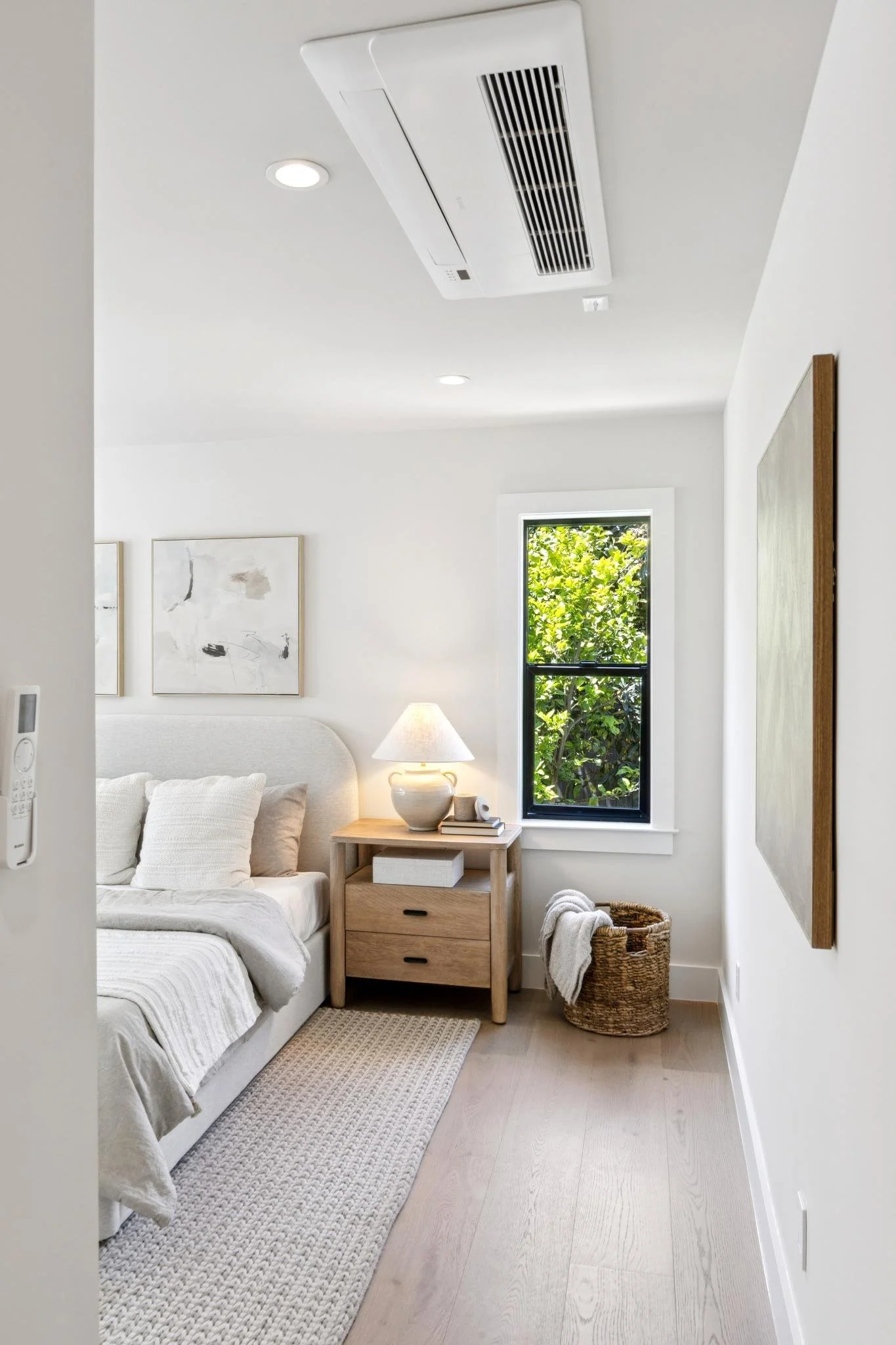 A cozy bedroom corner with a white bed, beige pillows, a small wooden nightstand with a lamp, a basket with a throw blanket, abstract wall art, a window with green foliage outside, and a ceiling air vent.