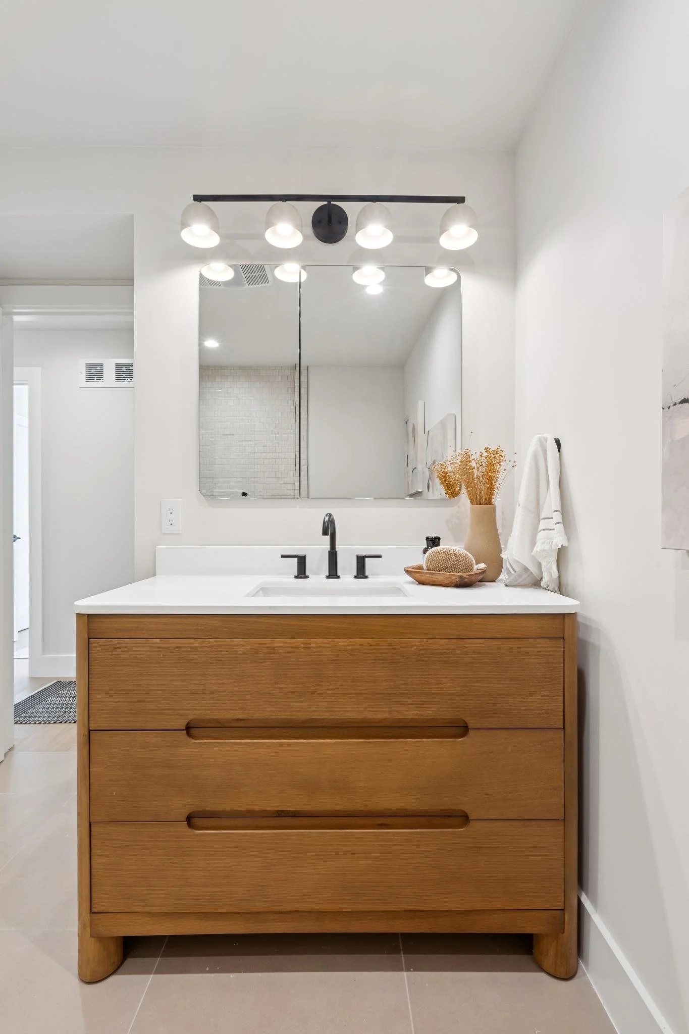 Modern bathroom vanity with a white countertop, black faucet, and a mirror above. Decorated with a vase of dried flowers, a round wooden container, and a white towel hanging on the side.