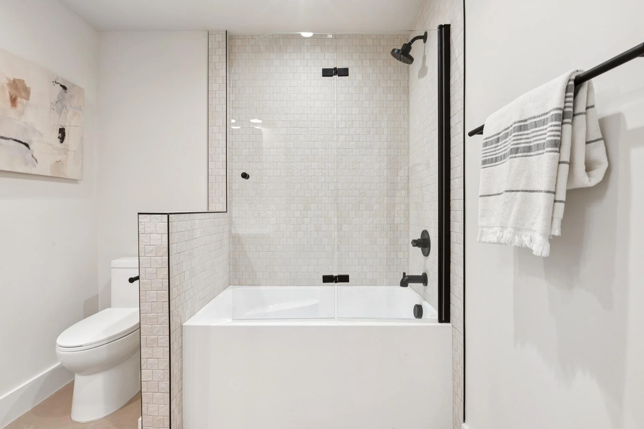 A modern bathroom featuring a walk-in shower with glass doors, black fixtures, white tiled walls, a white toilet, a towel on a black rail, and a piece of abstract art on the beige wall.