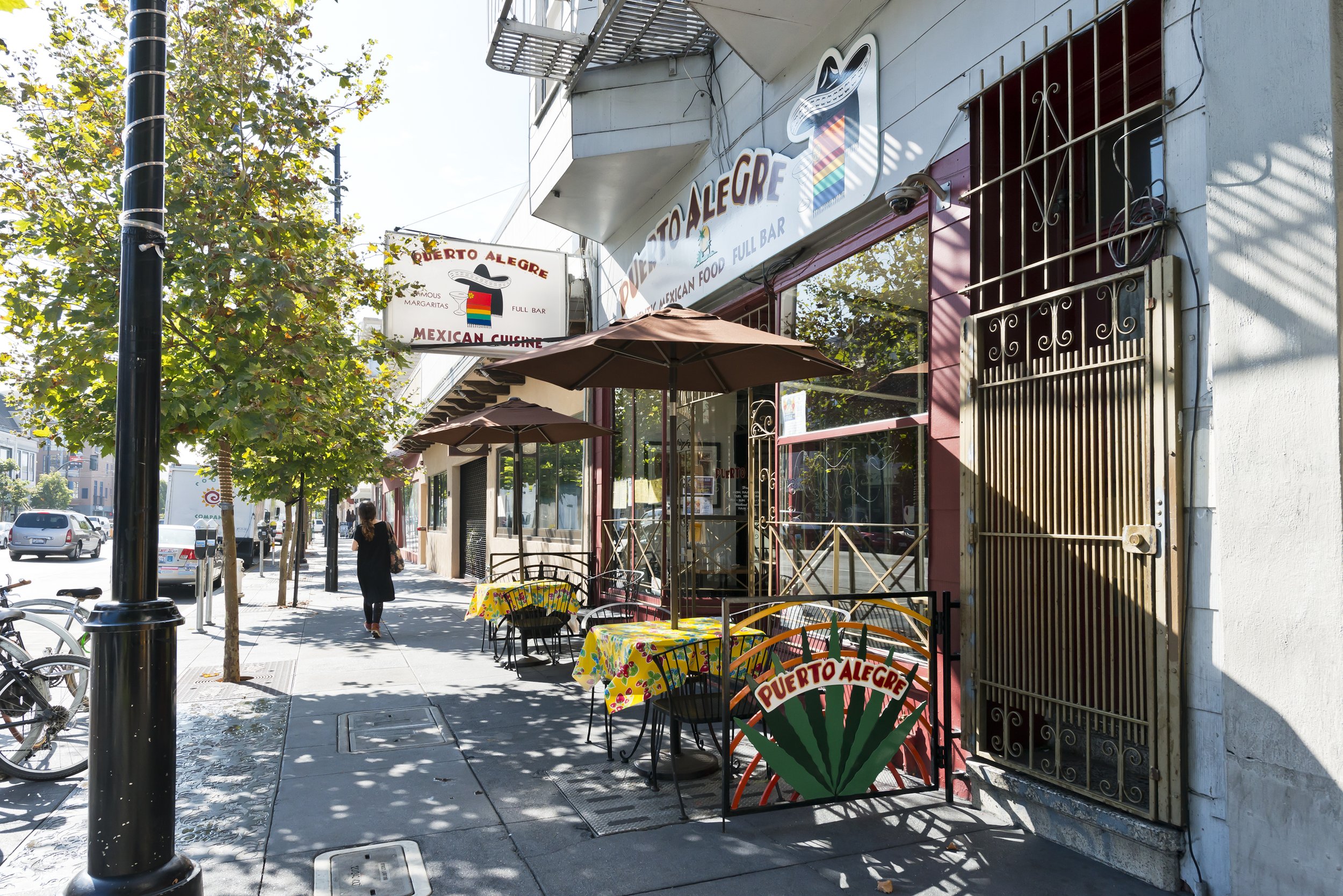 Street view of a Mexican restaurant called Puerta Alegre with outdoor seating on the sidewalk, colorful tables with tablecloths, umbrellas, trees, parked cars, and pedestrians walking by.