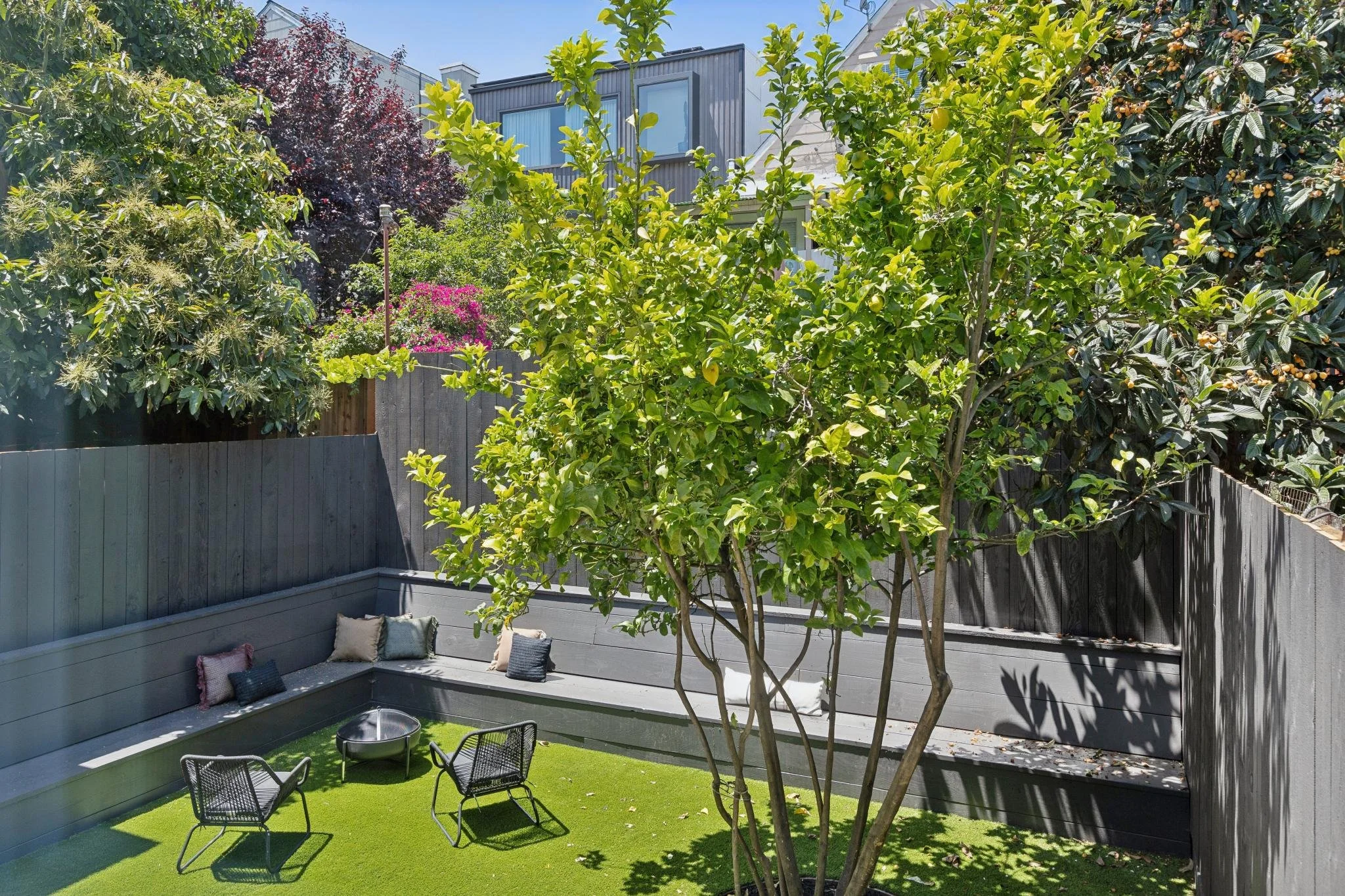 A backyard scene with a grey deck, garden chairs, a small garden bed, green grass, and various trees and plants under a clear blue sky.