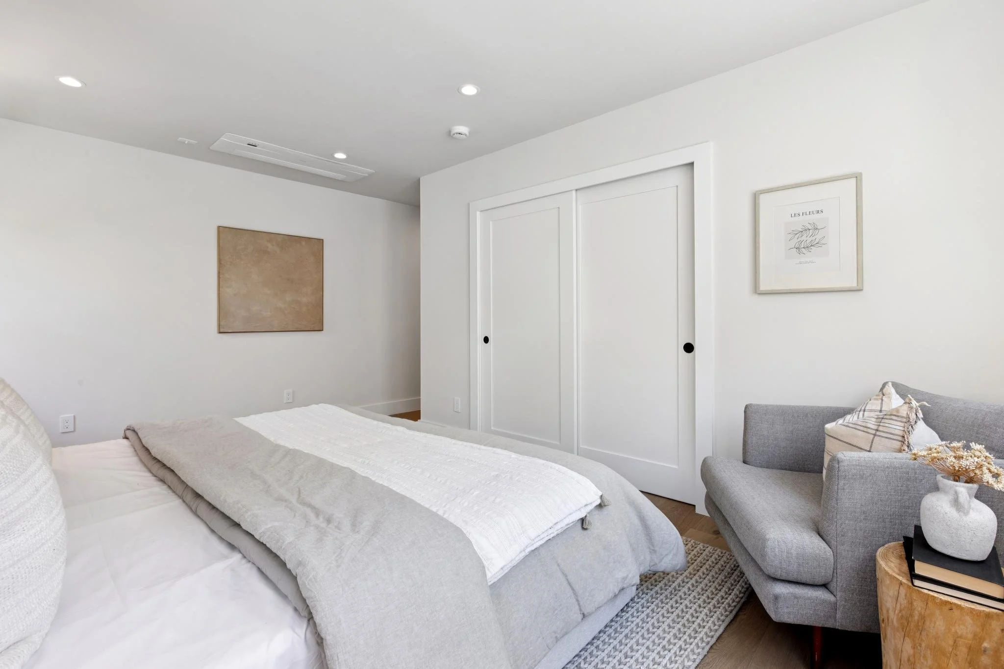 A bedroom with white walls, a bed with a white blanket, a gray armchair, framed wall art, and a closet with sliding doors.