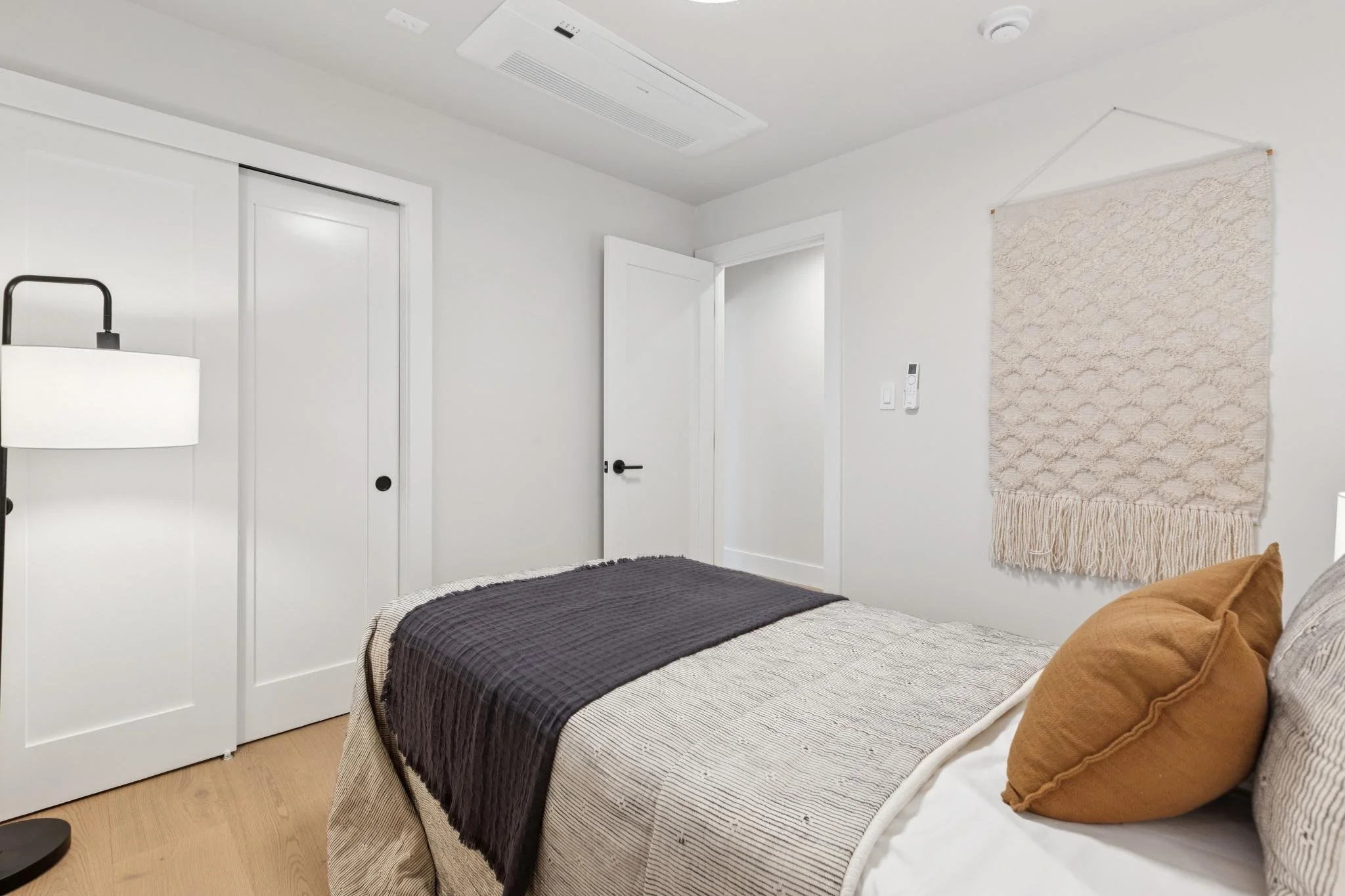 A bedroom with a bed, brown pillows, white bedding, a black blanket, a wall hanging, a door, built-in closet with sliding doors, a lamp, walls, and wooden flooring.