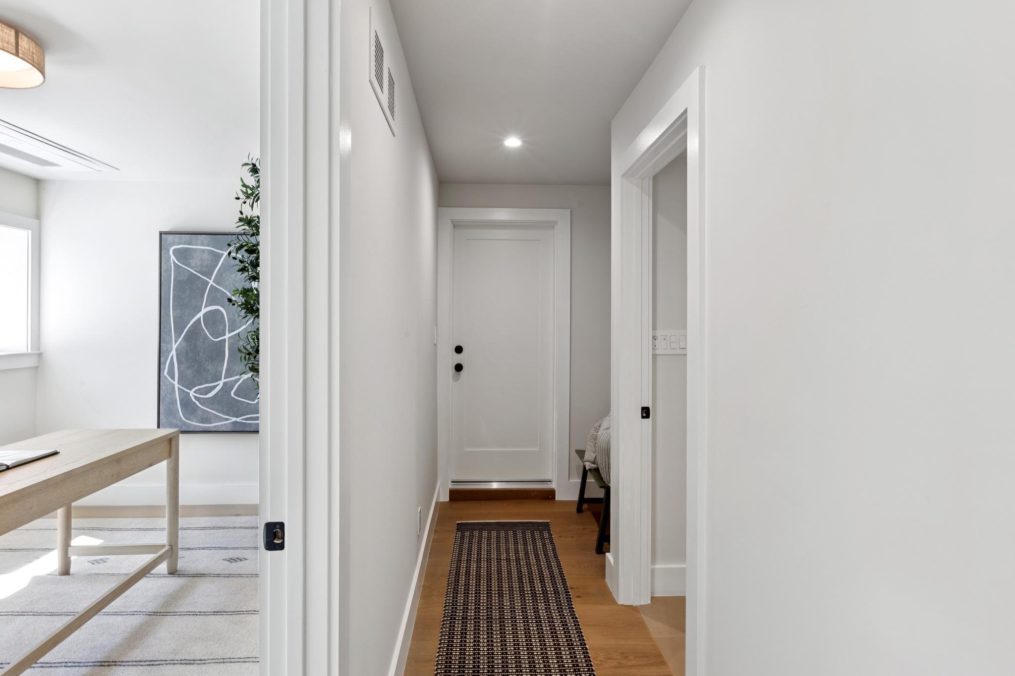 A narrow hallway with white walls, hardwood floor, and a small black and white striped rug. There is a white door at the end of the hallway and a small chair with a blanket on it on the right side. The hallway opens into a bright room with a piece of