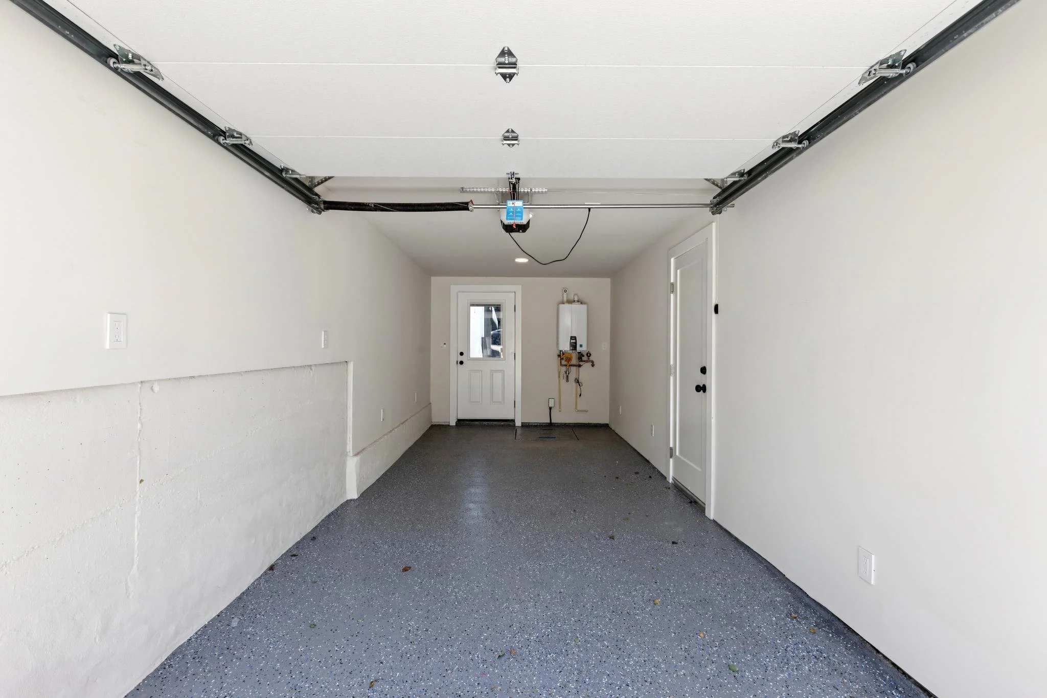 Empty garage with white walls, gray speckled floor, garage door opener installed on the ceiling, and a door at the back leading outside.